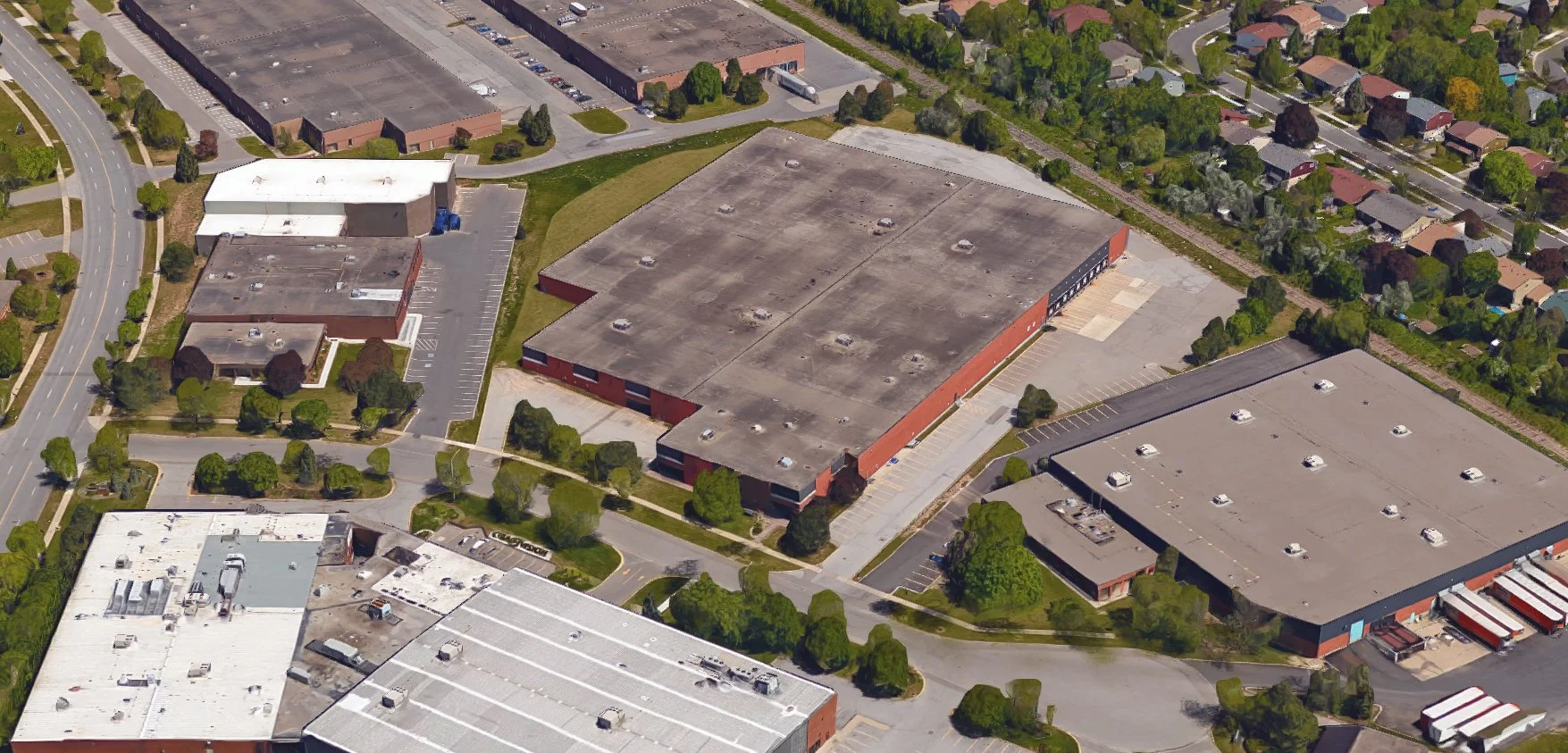 An aerial view of a commercial area with multiple large warehouse buildings, parking lots, and surrounding greenery, including trees and residential houses.