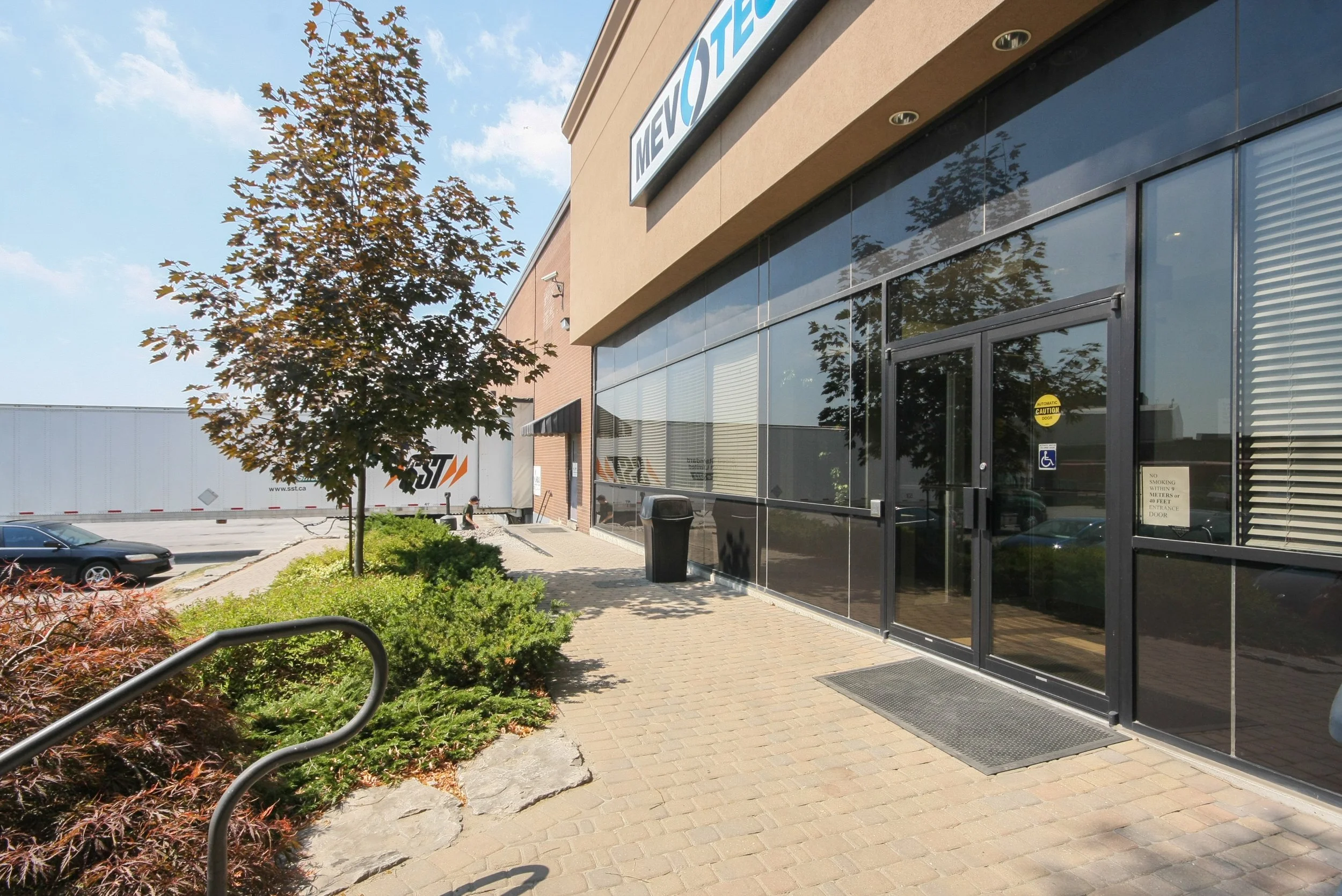 Exterior view of a modern commercial building with large glass windows and a sign that says 'METRO'. There is a tree with autumn-colored leaves, parked cars, and a truck in the background. The sidewalk has a textured surface and a ramp for accessibil