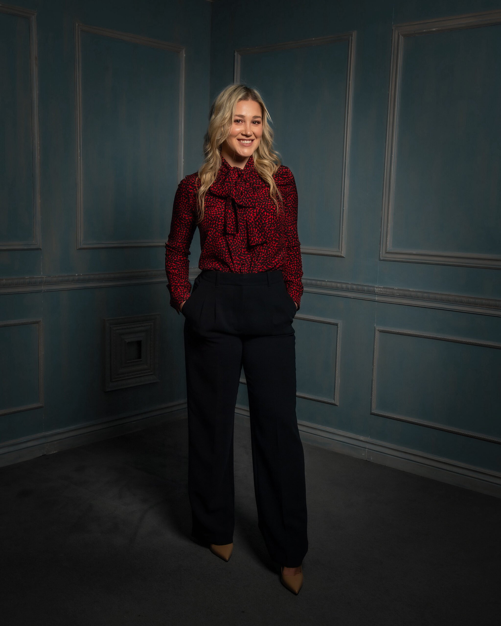 A woman with long blonde hair smiling, wearing a red and black patterned blouse with a bow tie at the collar and black pants, standing in a darkened room with teal walls decorated with rectangular molding.