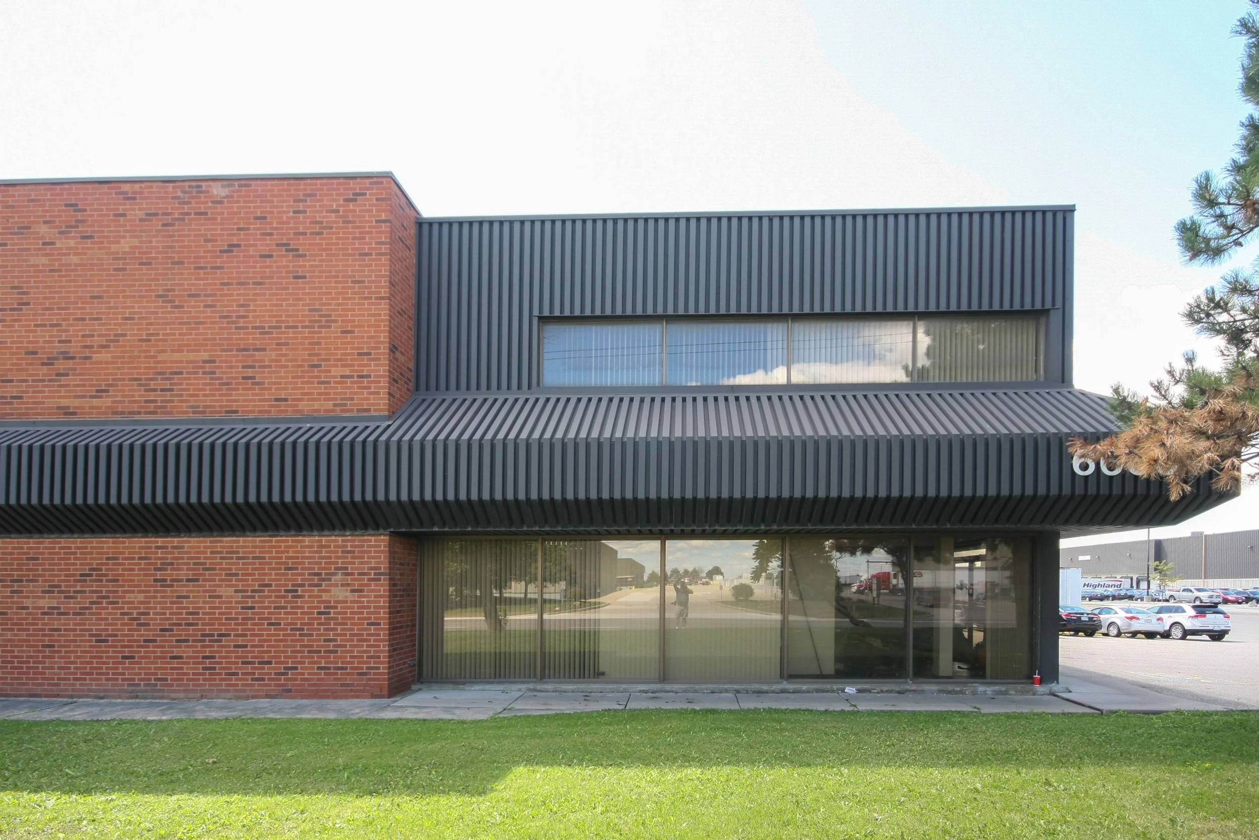 Modern commercial building with red brick wall and black metal panel accents, large windows reflecting the sky, and a grassy lawn in the foreground.