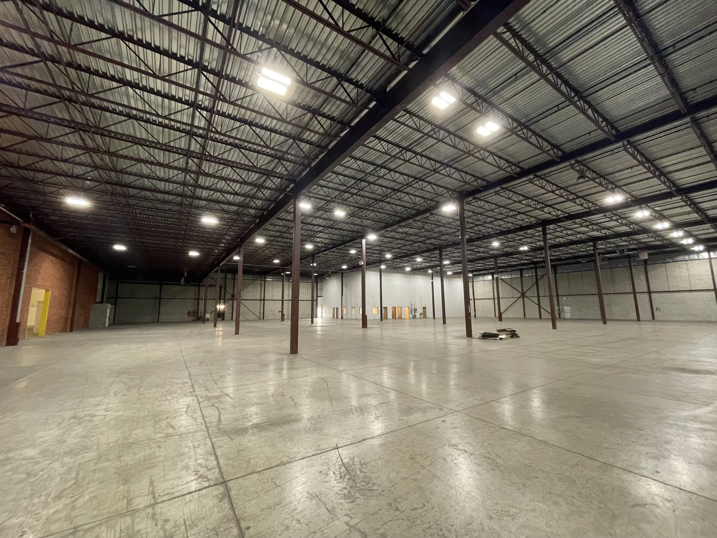 Interior of a large, empty warehouse with concrete floors and metal roof supported by steel beams, illuminated by ceiling lights.