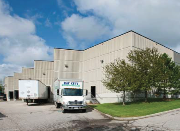 A warehouse with trucks parked outside, surrounded by a grassy area and trees under a partly cloudy sky.
