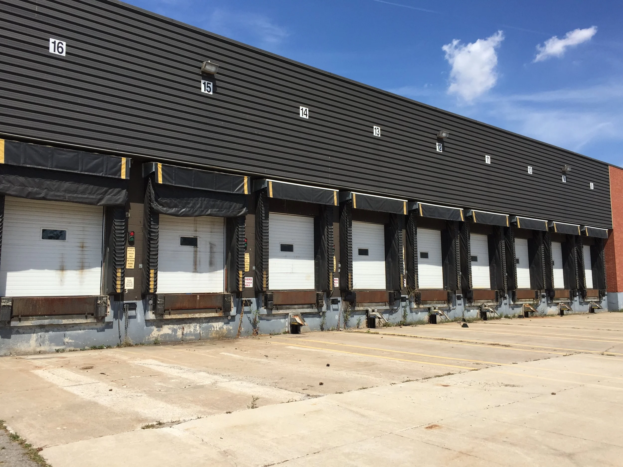 An empty parking lot in front of a commercial building with closed garage doors. The building has a black metal facade with numbered sections and exterior lights. The sky is partly cloudy with blue and white clouds.