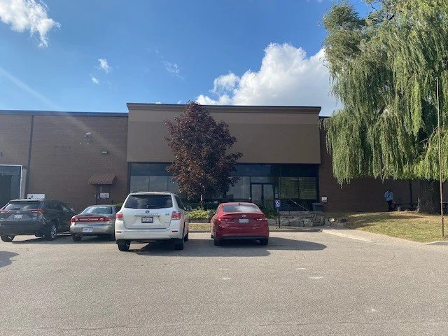 Parking lot outside a commercial building with several parked cars, a tree in front, and a blue sky with clouds.