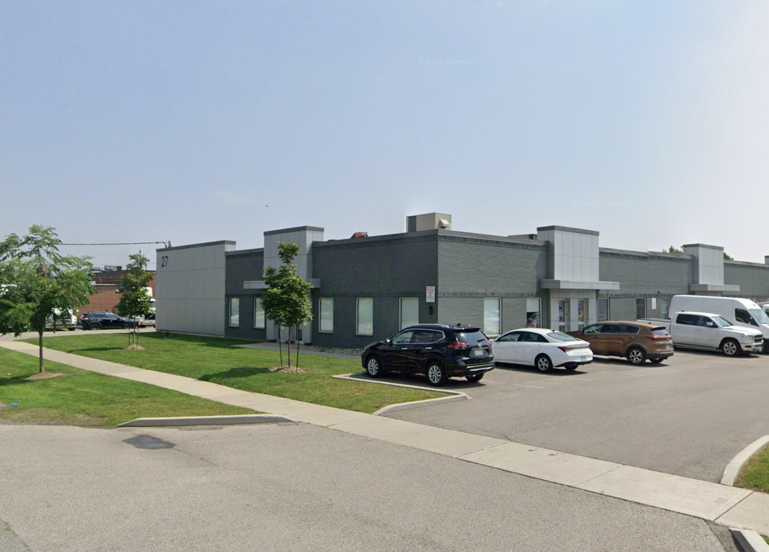 A modern commercial or office building with a parking lot in front, several parked cars, and some young trees with green leaves. The sky is clear and blue.