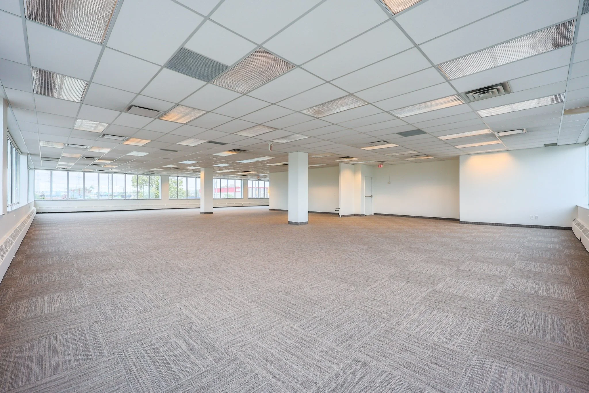 Empty office space with large windows, white walls, a tiled suspended ceiling, and beige carpeted floor.