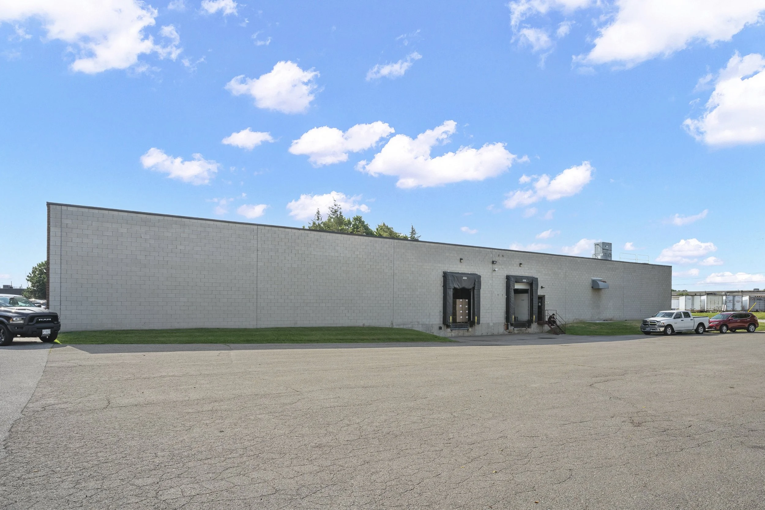 Large beige brick building with loading docks, parked cars, and a small patch of grass in the foreground under a blue sky with white clouds.