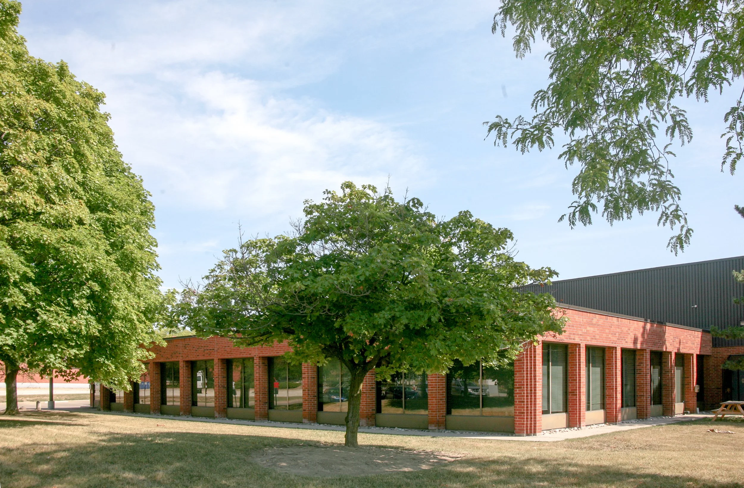 A brick building with large windows and a dark roof surrounded by green trees and a grassy area, with a blue sky overhead.