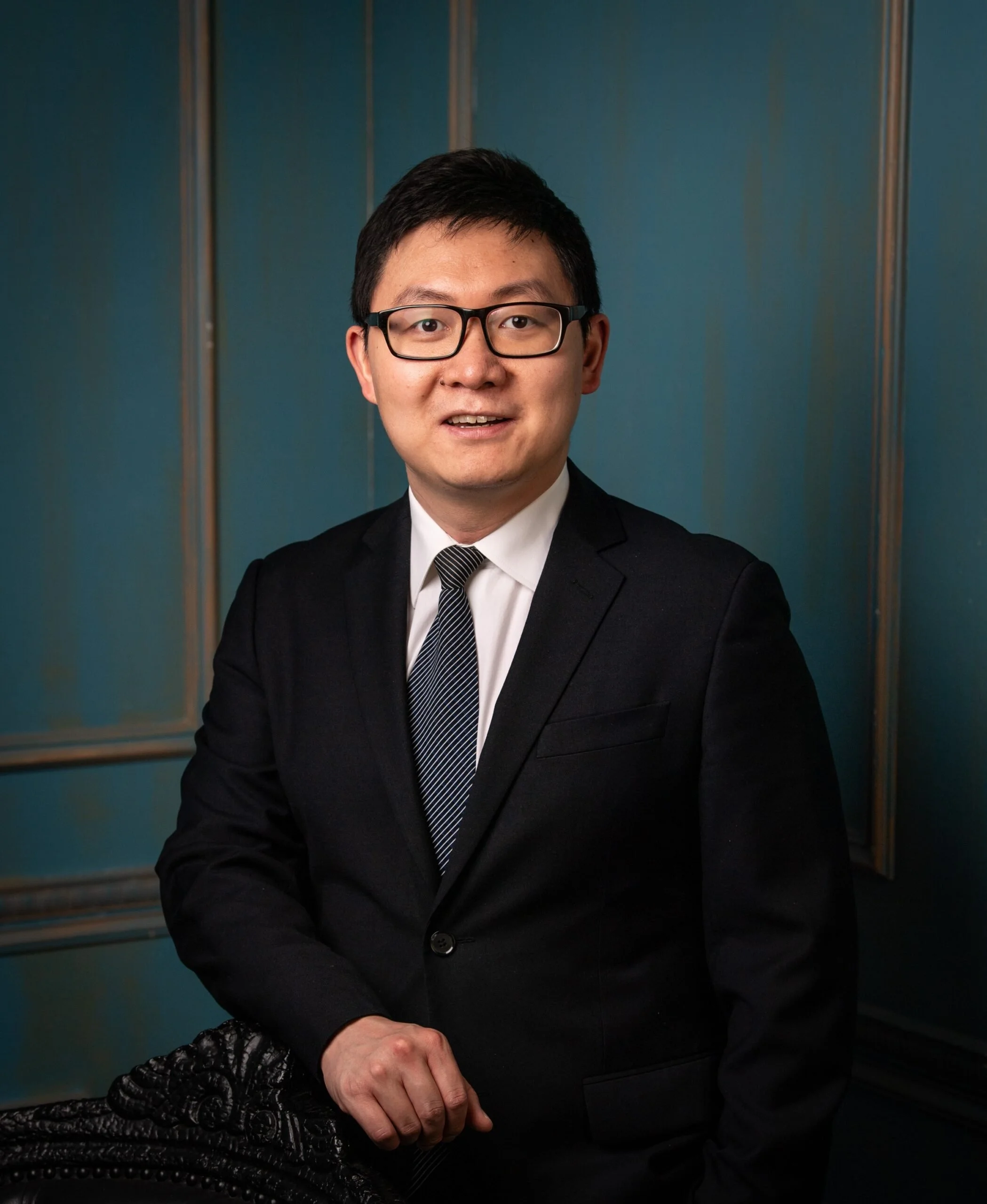A man wearing glasses, dressed in a black suit, white shirt, and striped tie, standing in front of a teal wall with vertical metallic accents.