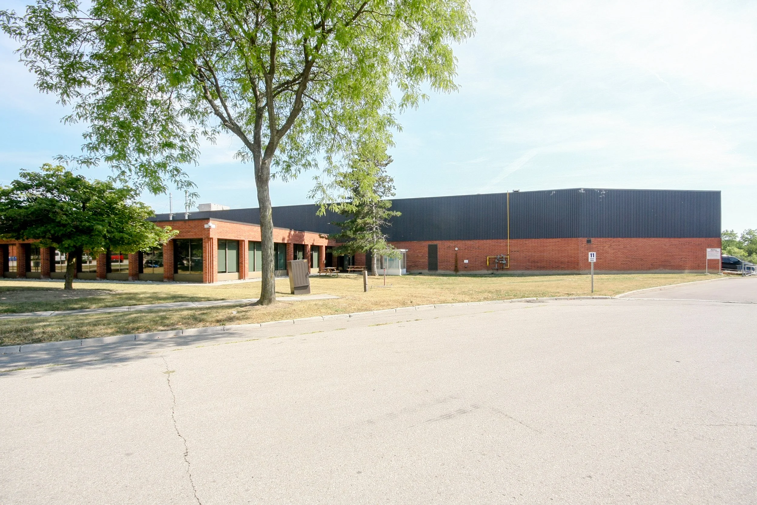 A large building with red brick walls and a black roof, surrounded by green trees, with a parking lot and a street in the foreground.