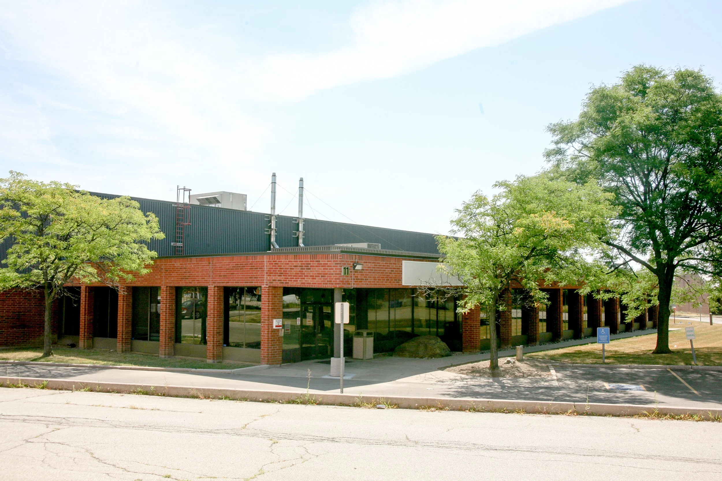A single-story modern building with large glass windows and brick columns at the entrance, surrounded by green trees and a parking lot, under a clear blue sky.