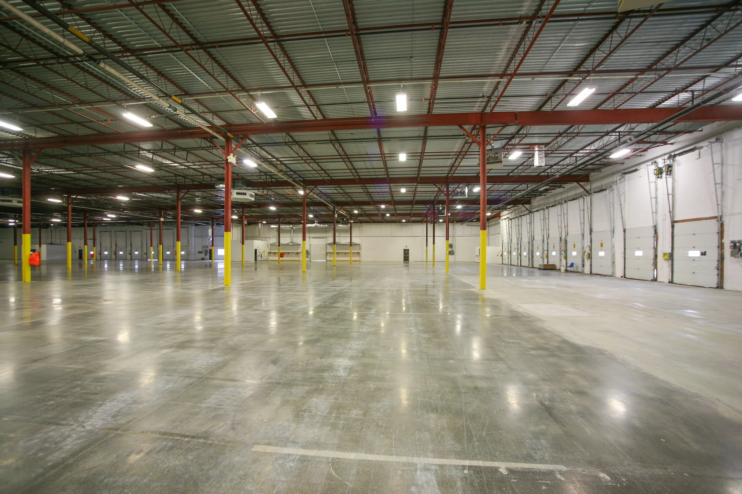 Large empty warehouse with a high ceiling, red steel beams, and bright overhead lighting.