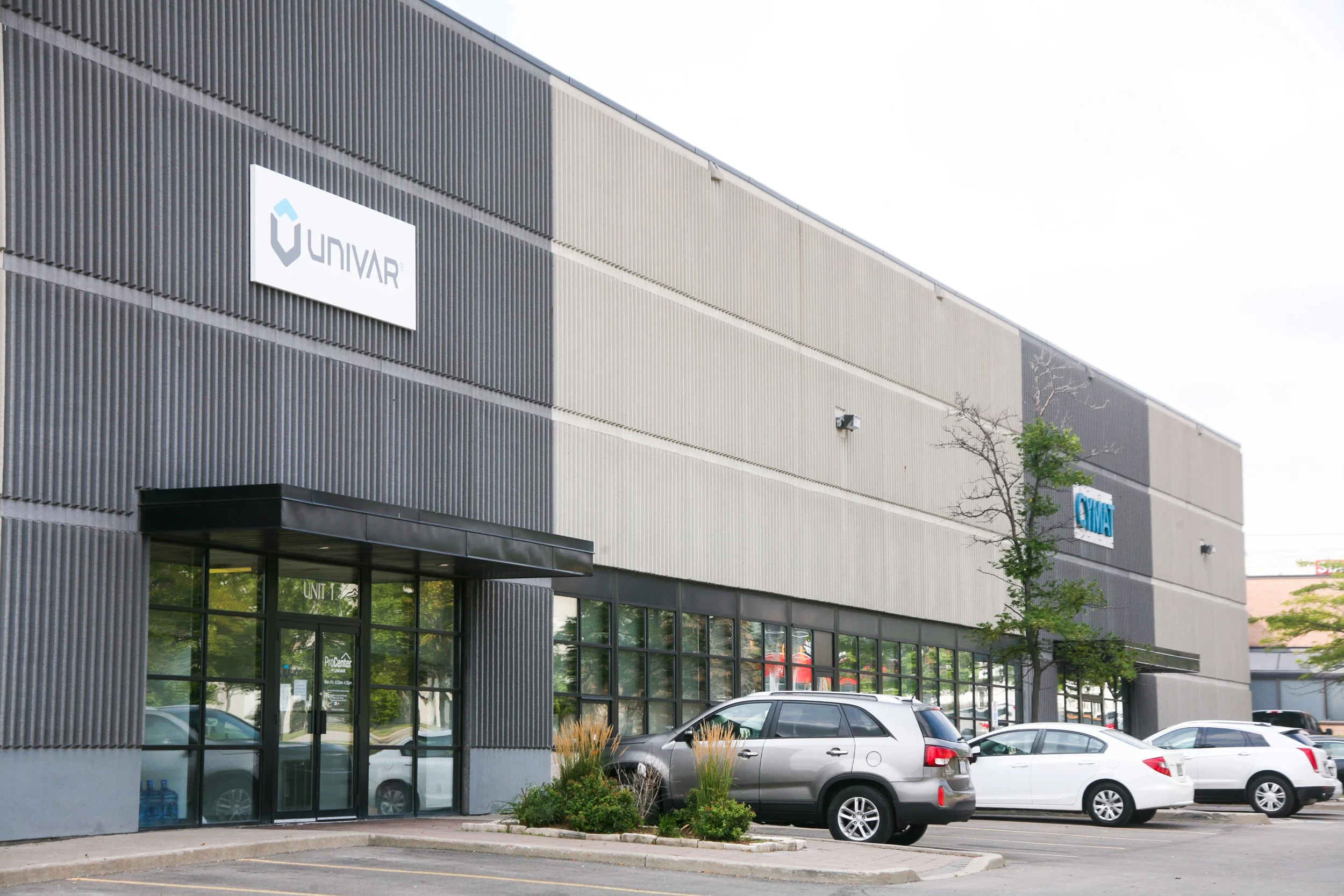 Exterior view of a commercial building with a sign that reads 'UNIVAR'. The building has a modern design with a textured gray facade and large windows. There are parked cars and a small landscaped area with plants and trees in the parking lot.
