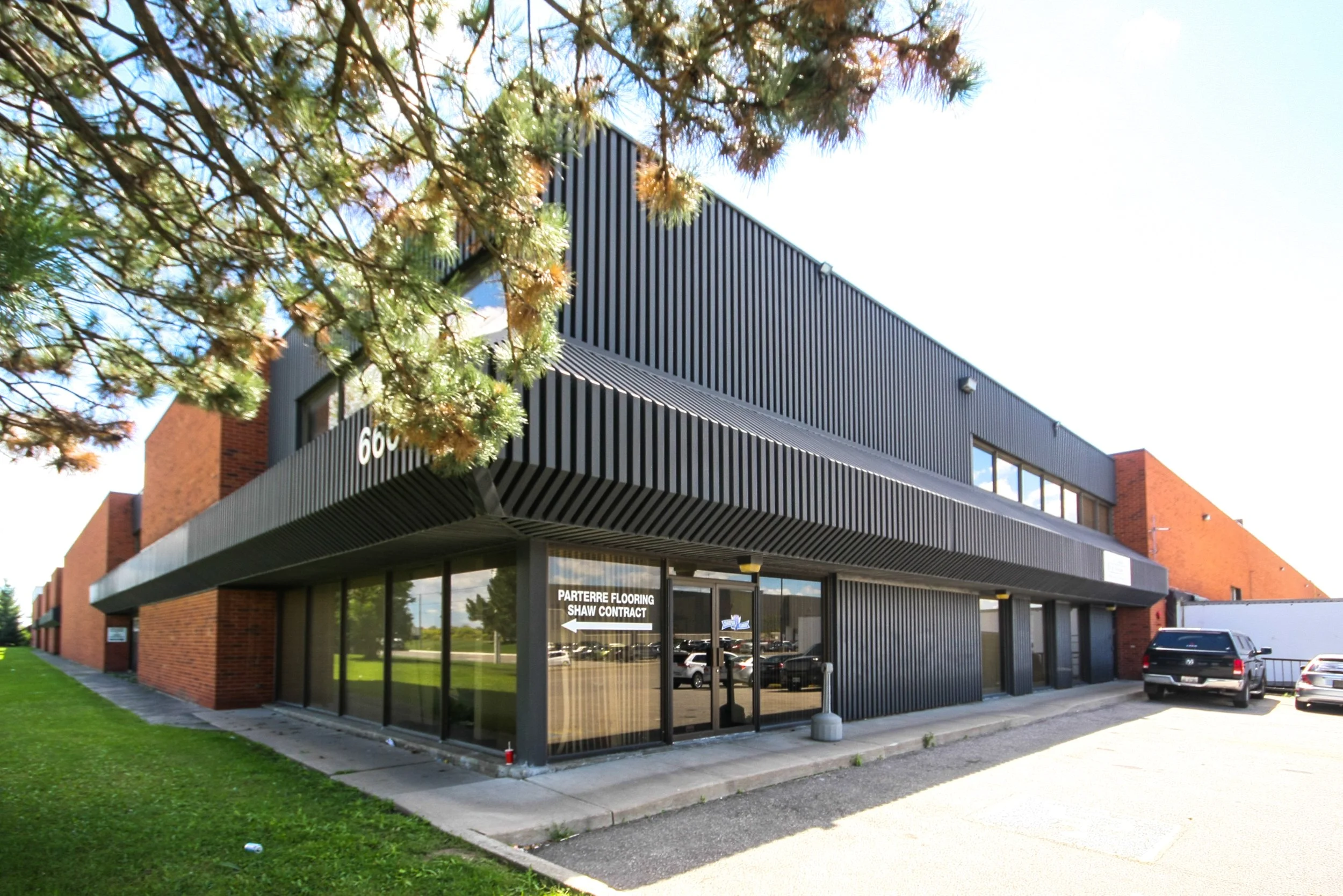 Modern two-story commercial building with black metal siding, large glass windows, and brick accents, situated on a paved lot with parked cars. A tree with green and brown leaves partially obscures the view, and a sign points to flooring and contract