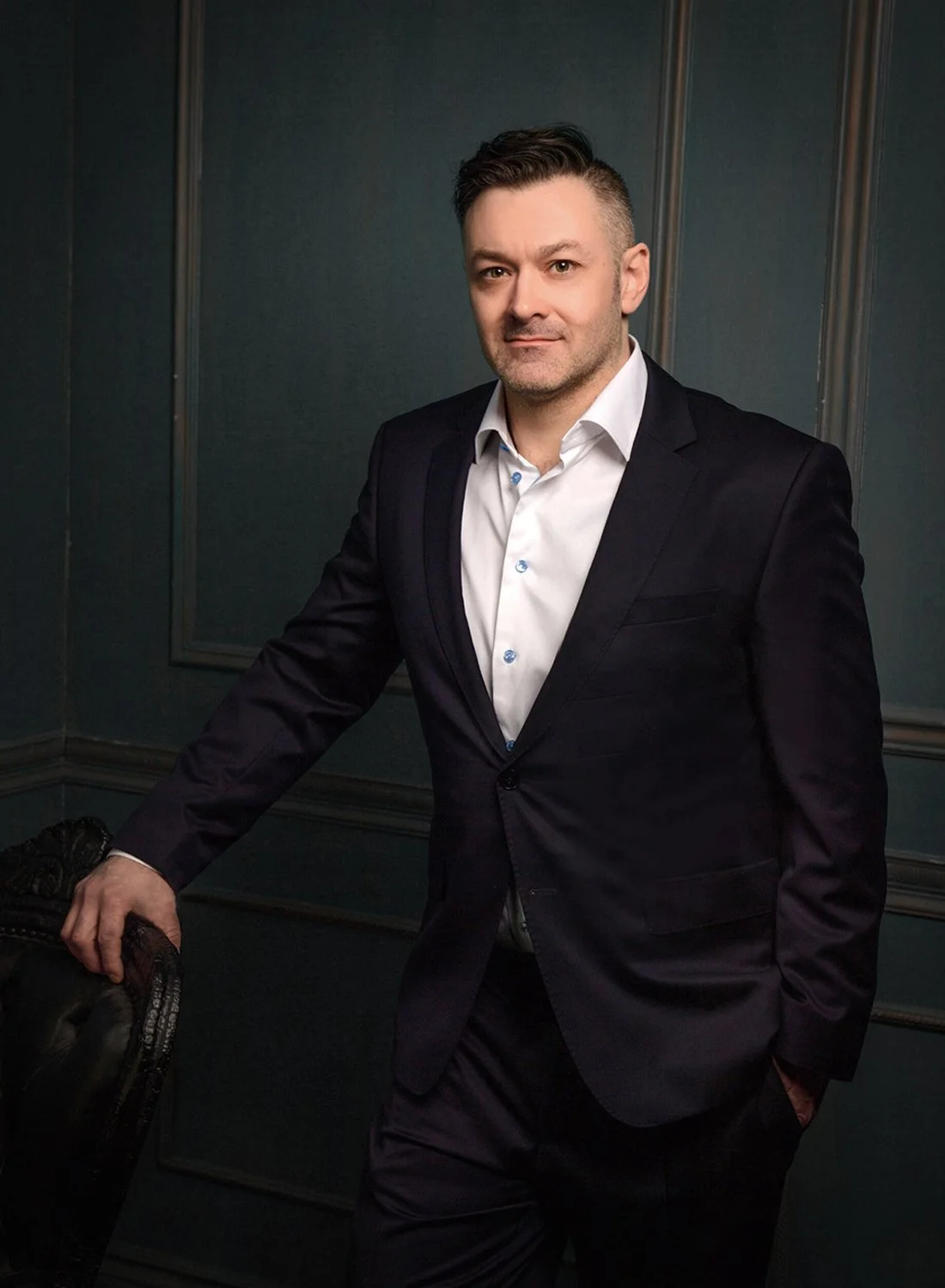 A man in a dark suit and white shirt standing in a room with dark green walls, leaning on a chair, looking at the camera.