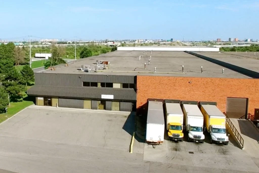 An industrial building with three delivery trucks parked outside on a paved lot, with a large flat roof and a tree-lined area nearby.