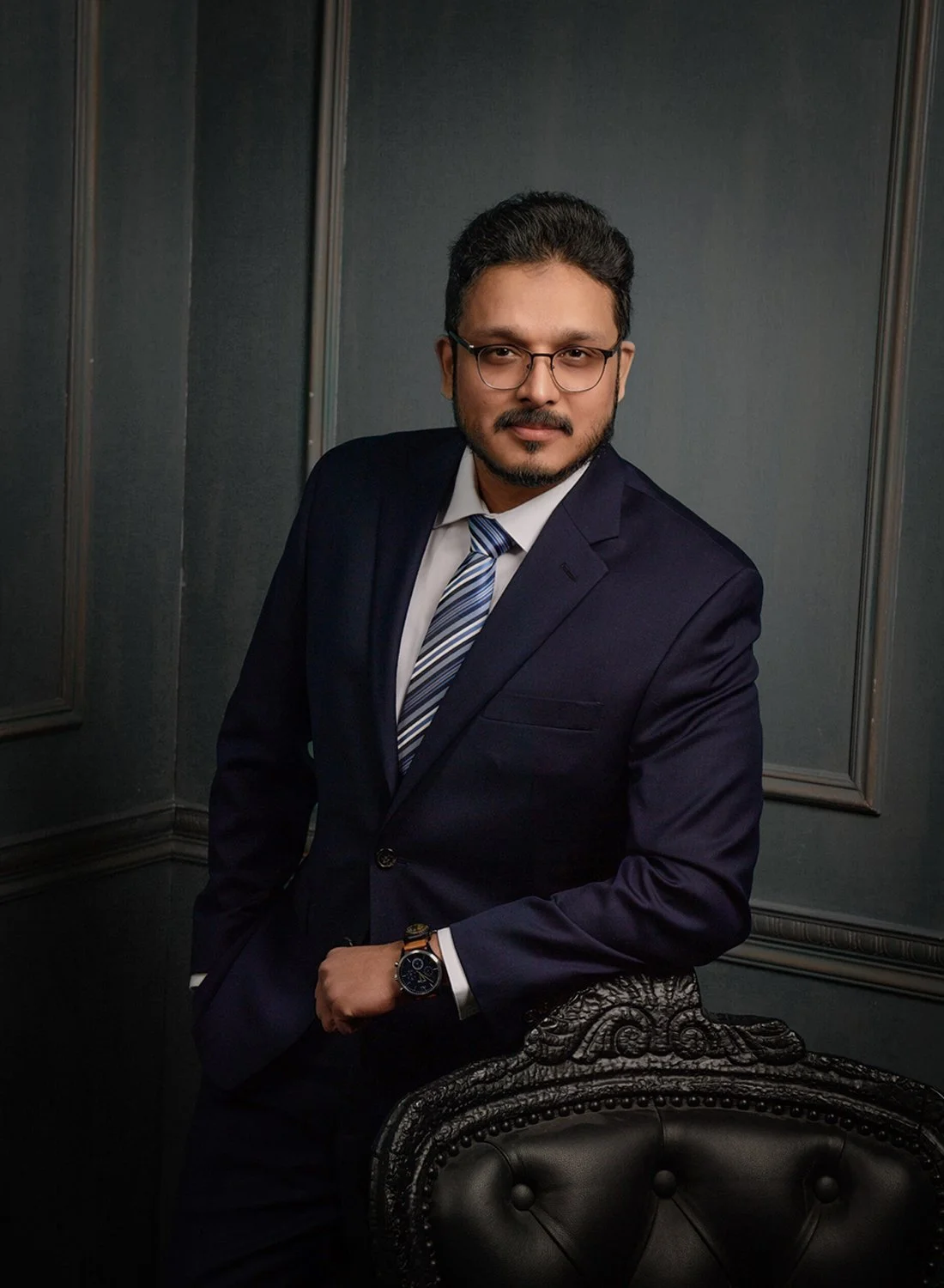 A man in a dark suit, white shirt, and striped tie, wearing glasses and a watch, standing in a dimly lit room with dark walls and ornate furniture.
