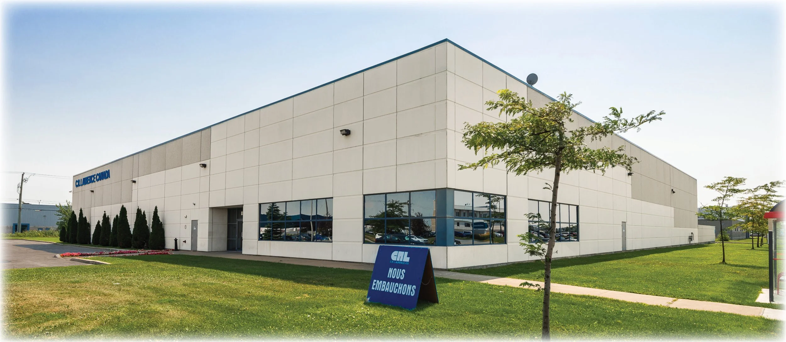 Large white industrial building with blue signage and windows, surrounded by green grass and trees, sunny sky, parking lot reflections visible in windows, sign outside reading 'Nous Embauchons' indicating they are hiring.