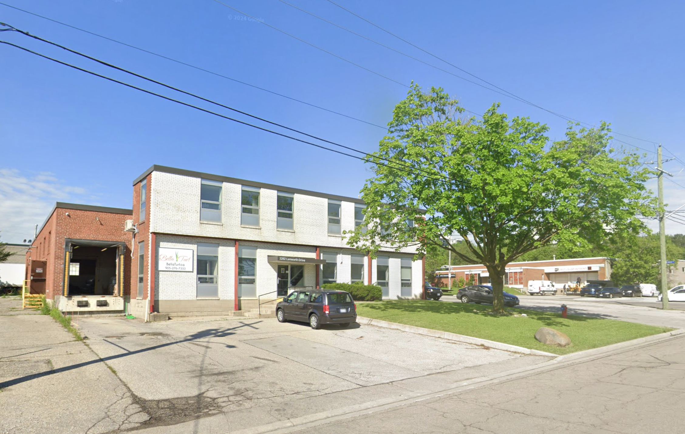 A city street view featuring a two-story brick and white building with signs and several parked cars, large green tree on the sidewalk, and utility poles with power lines under a bright blue sky.