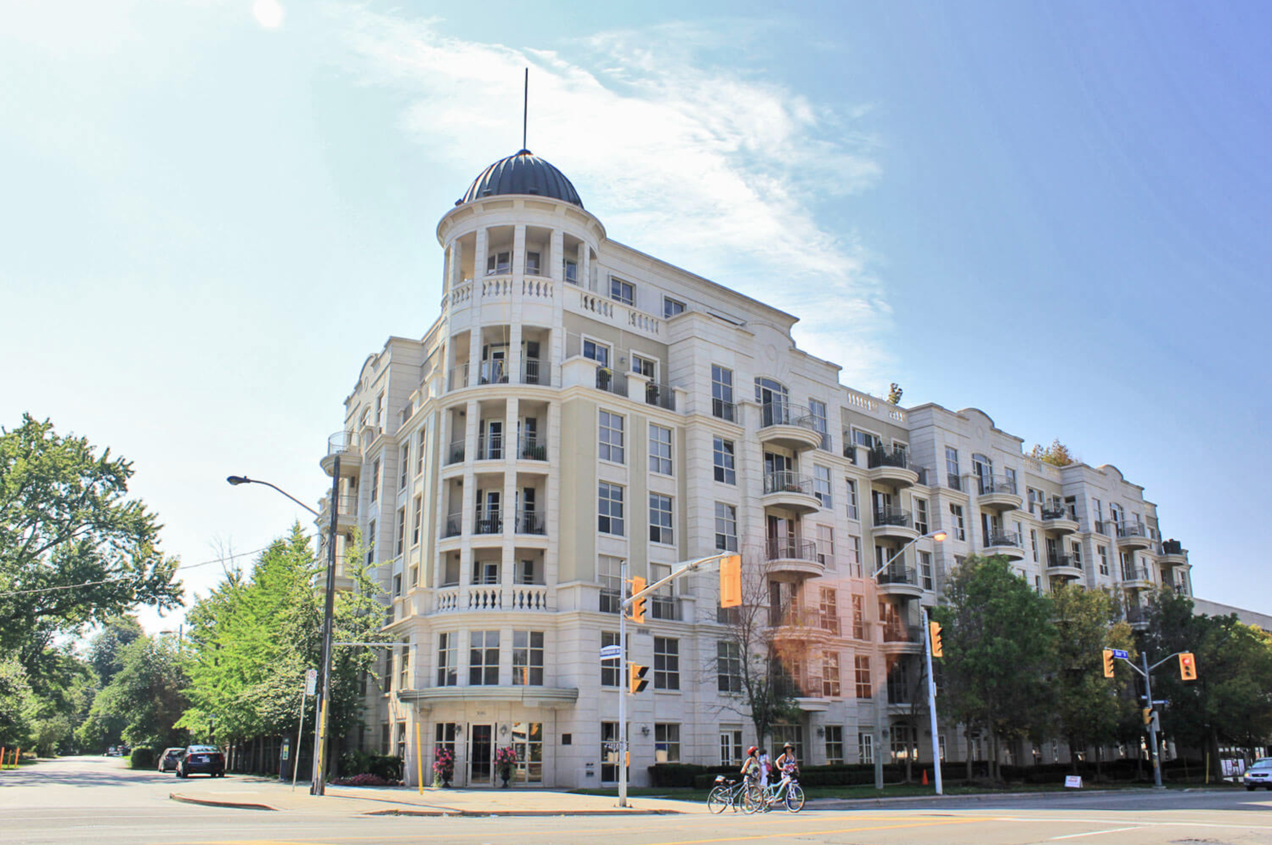 A tall, white, multi-story residential building with rounded balconies and a domed rooftop, situated at an intersection with traffic lights and street signs, surrounded by green trees and a few cyclists on the street.