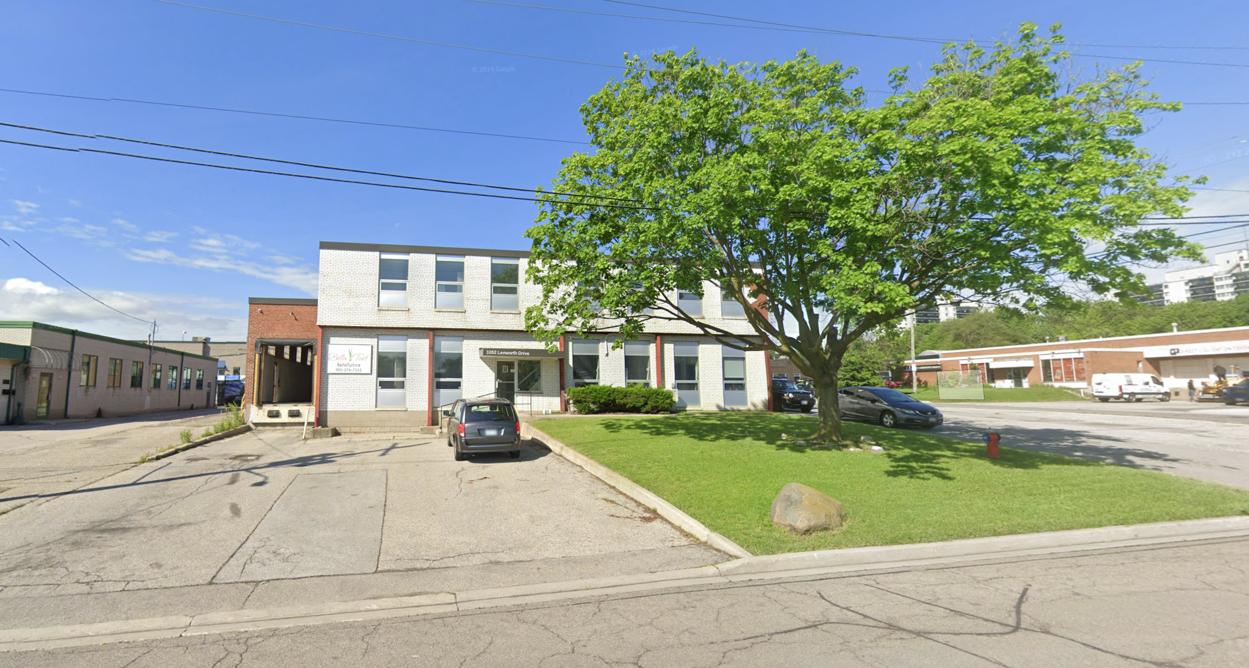 A parking lot with a few cars in front of a two-story building with white and brick walls, a large tree providing shade, and a clear blue sky with a few clouds.