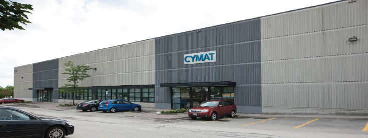 Exterior view of a large industrial building with a sign that says "CYMAT" and a parking lot with several cars.