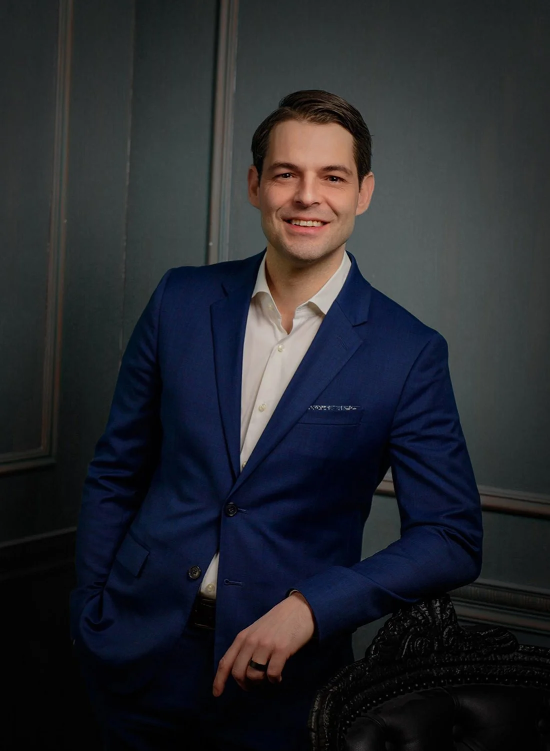 A man in a blue suit and white shirt smiling, standing in front of a dark wall.