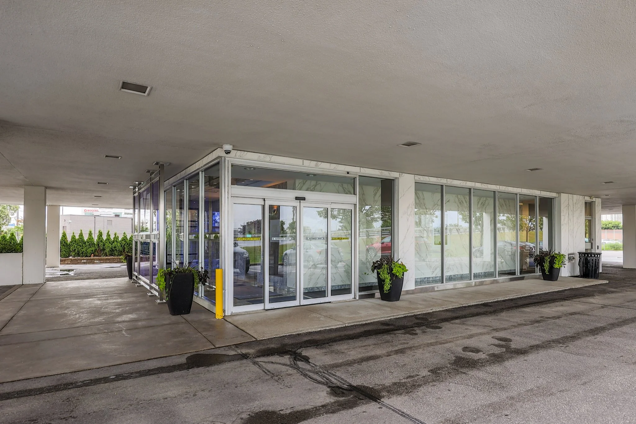 Exterior of a modern building with glass doors and marble walls, surrounded by potted plants. There is a parking lot and trees in the background.