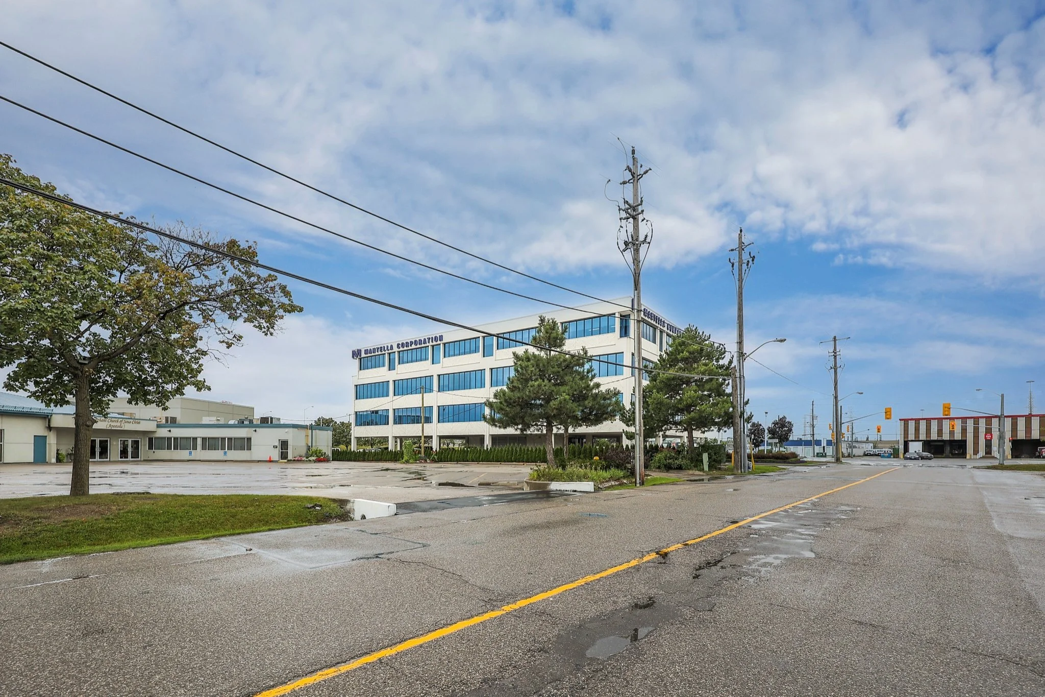 Empty parking lot with wet pavement and puddles, green trees, a white office building with blue windows labeled "Mantella Corporation," utility poles with power lines, and a partly cloudy sky.