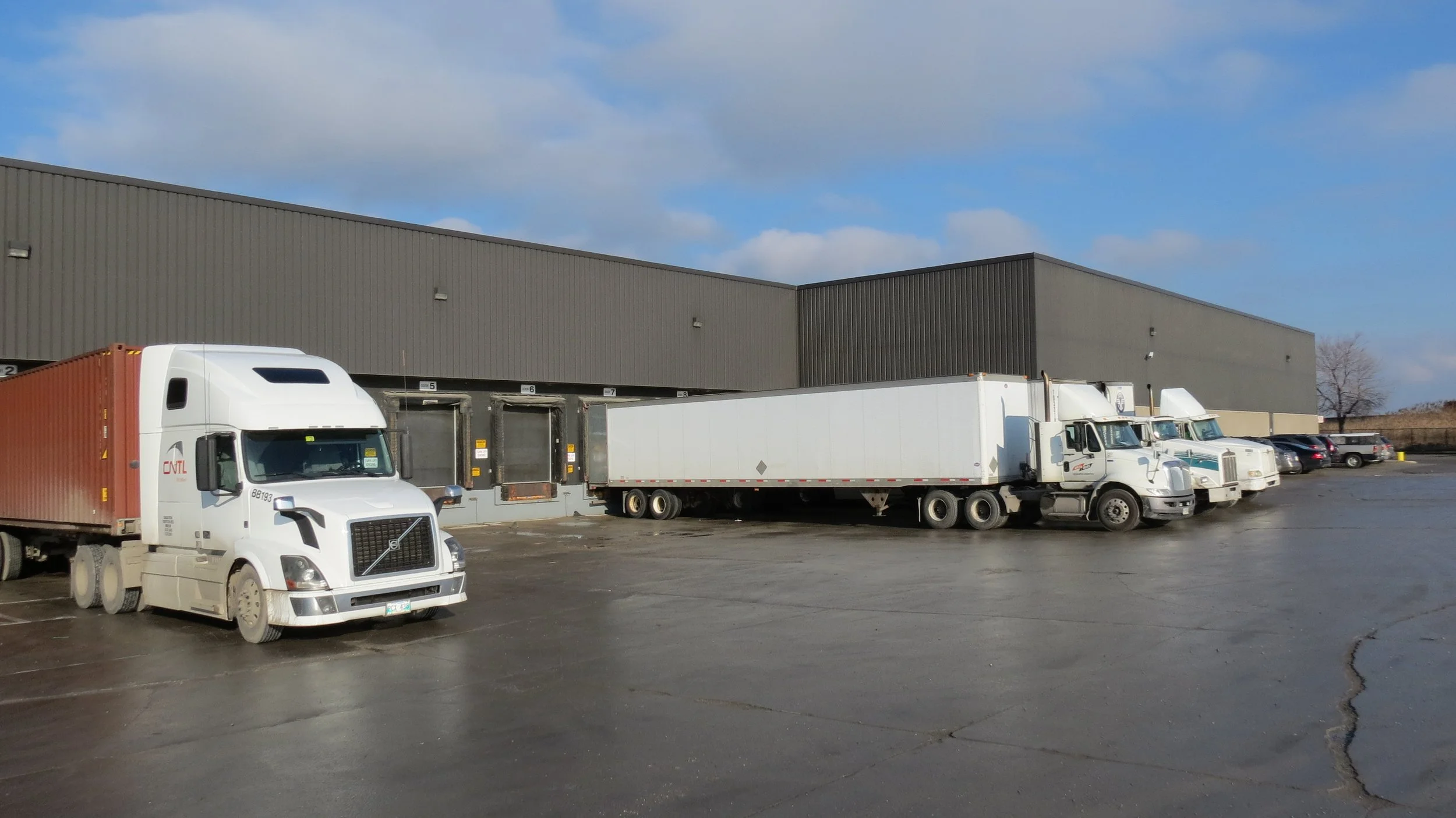 Parking lot in front of a large industrial warehouse with several trucks and cars parked.