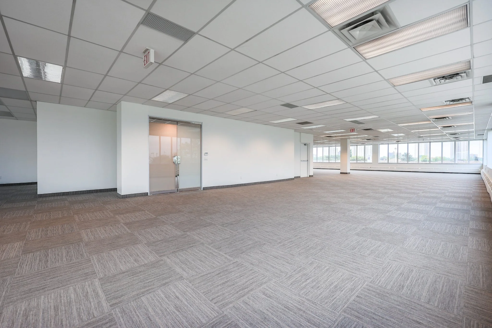 An empty office space with a drop ceiling, large windows, and carpeted flooring.