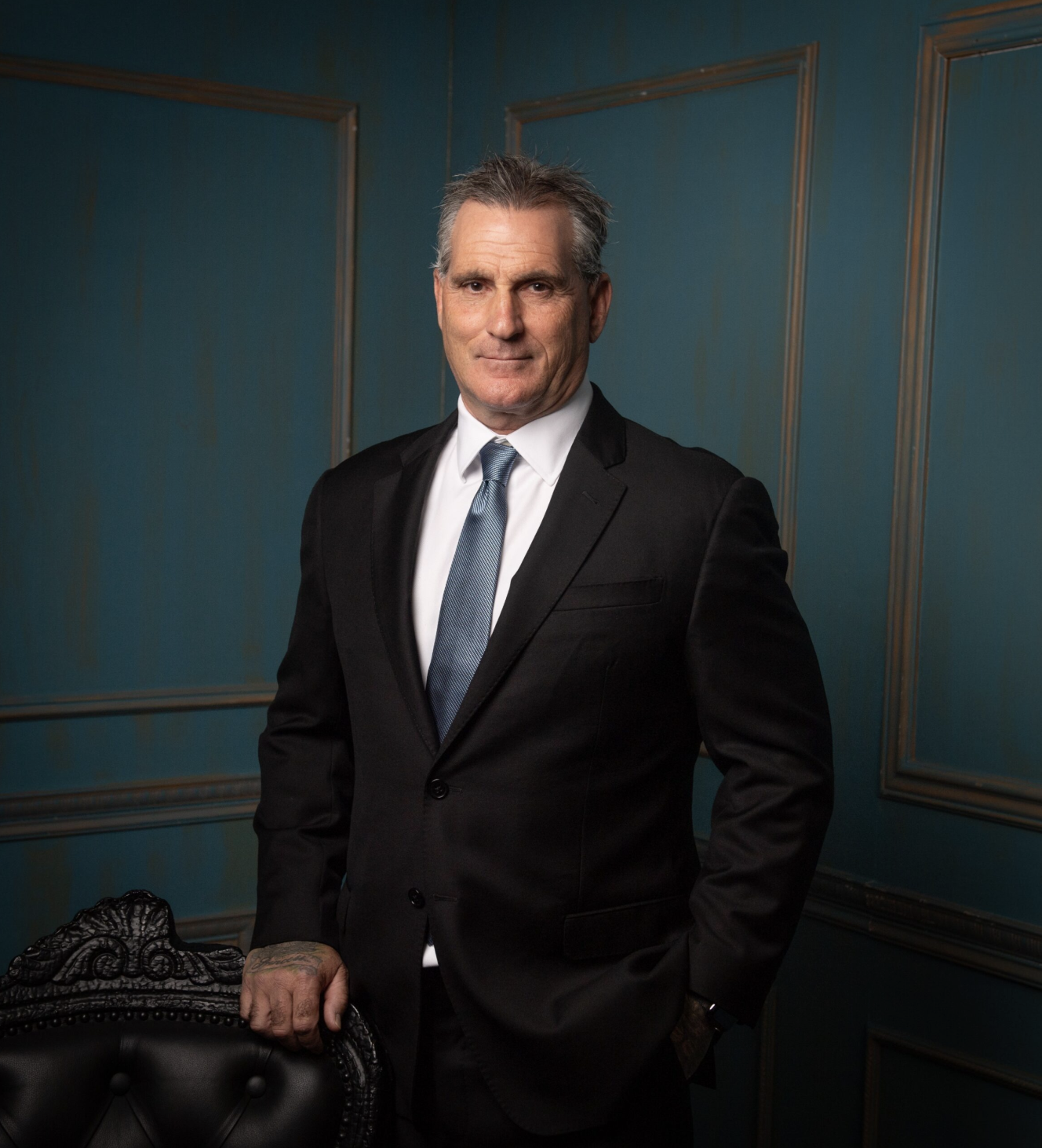 A man in a black suit and blue tie standing in a room with teal walls and decorative molding.