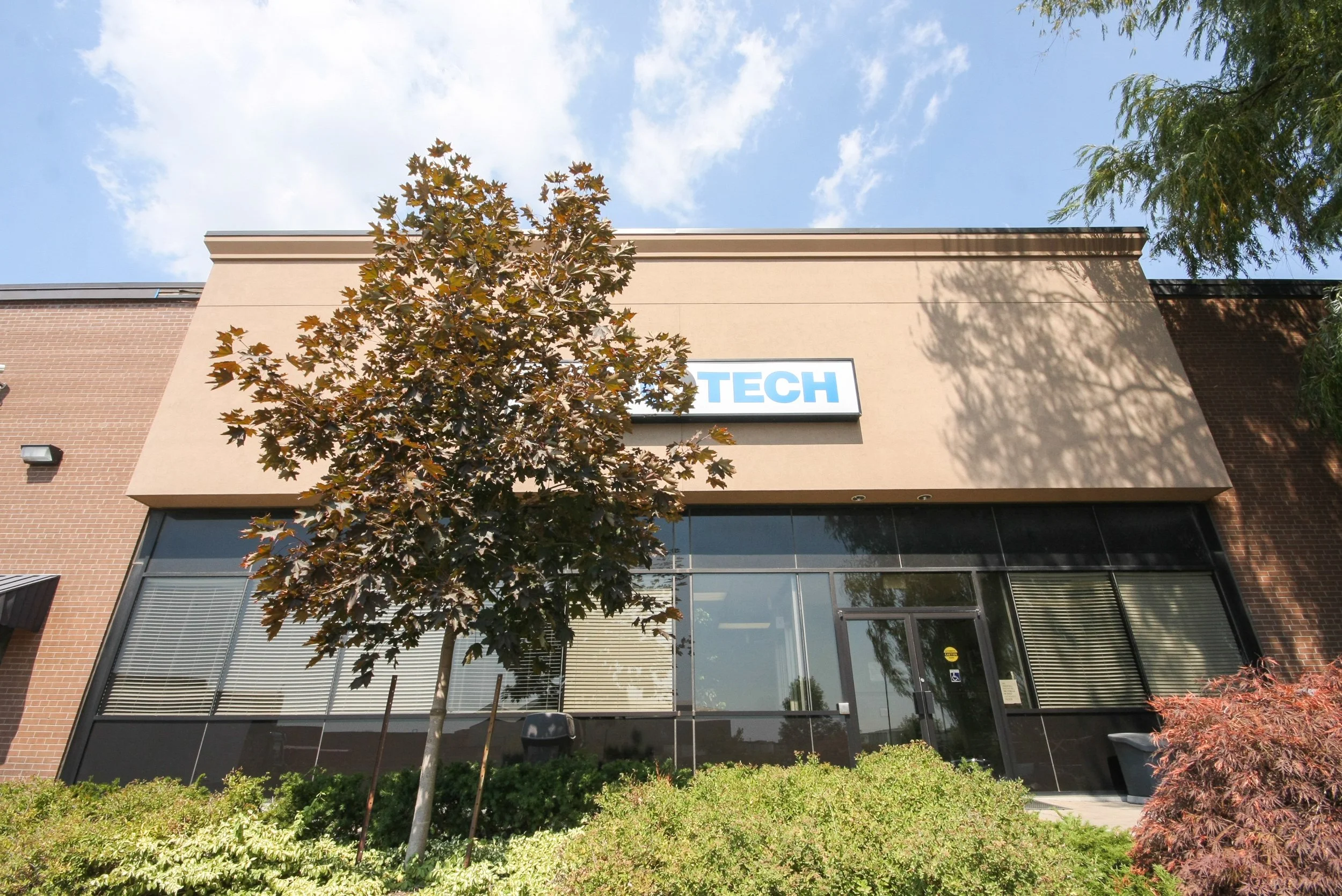 A modern commercial building with large windows and a brick facade, featuring a sign that says 'TECH', with trees and greenery in front, under a partly cloudy sky.