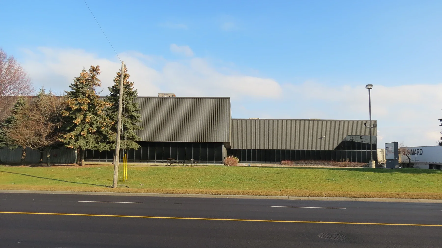 A large commercial building with a gray metal exterior, surrounded by trees and green grass. There are picnic tables, a streetlamp, utility pole, and a truck with the logo 'Smar' on the side in front of the building. The sky is mostly clear with a few clouds.