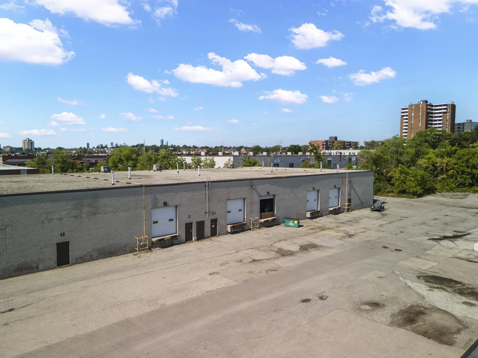 An industrial building in a parking lot with a city skyline and trees in the background under a partly cloudy sky.