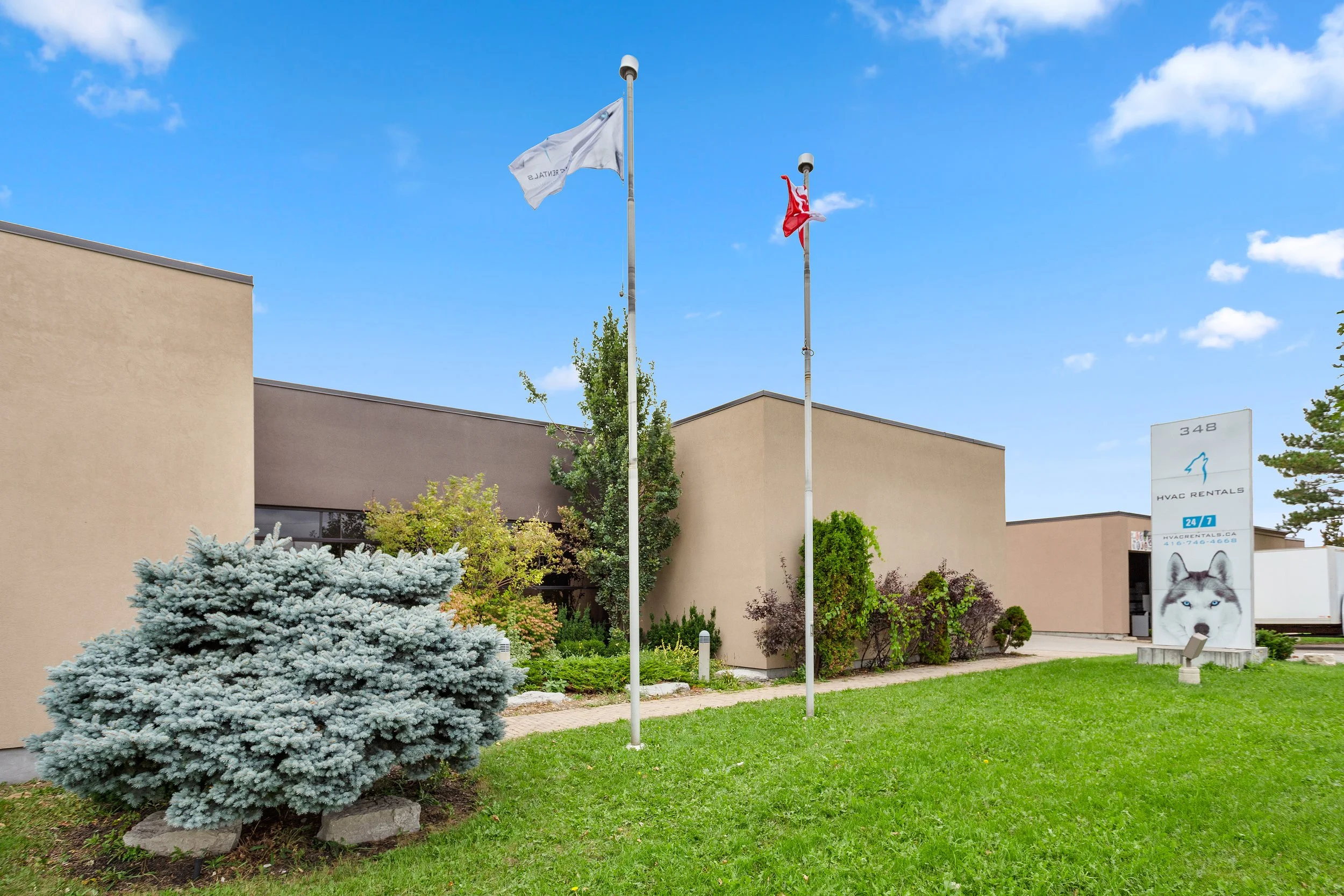 Two flagpoles with flags in front of a beige building, surrounded by landscaped greenery and trees, under a partly cloudy blue sky.