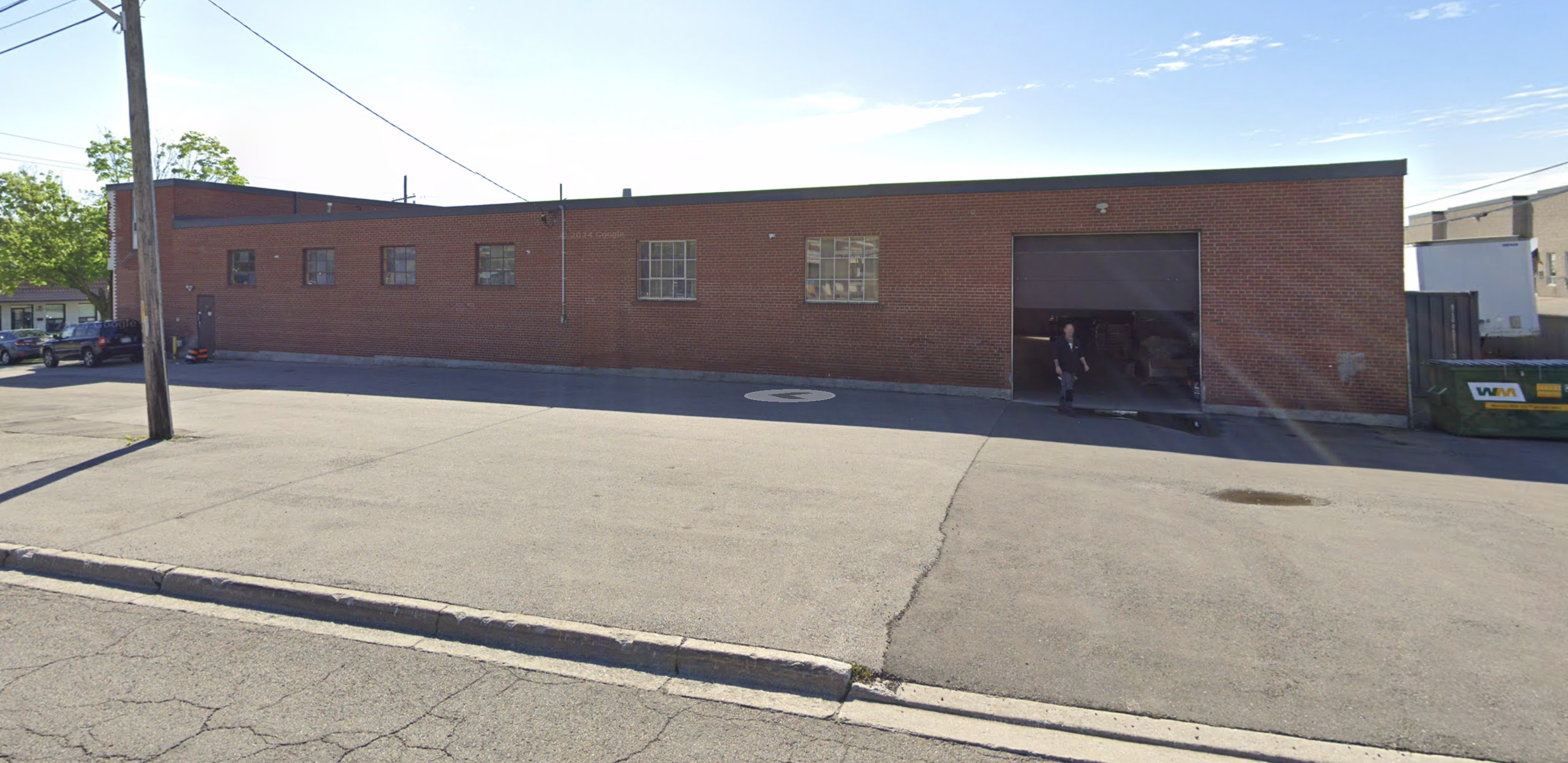 A brick building with a large garage door partially open, a man in a suit exiting, and several parked cars in front. There is a telephone pole on the left side of the image and a green dumpster on the right.