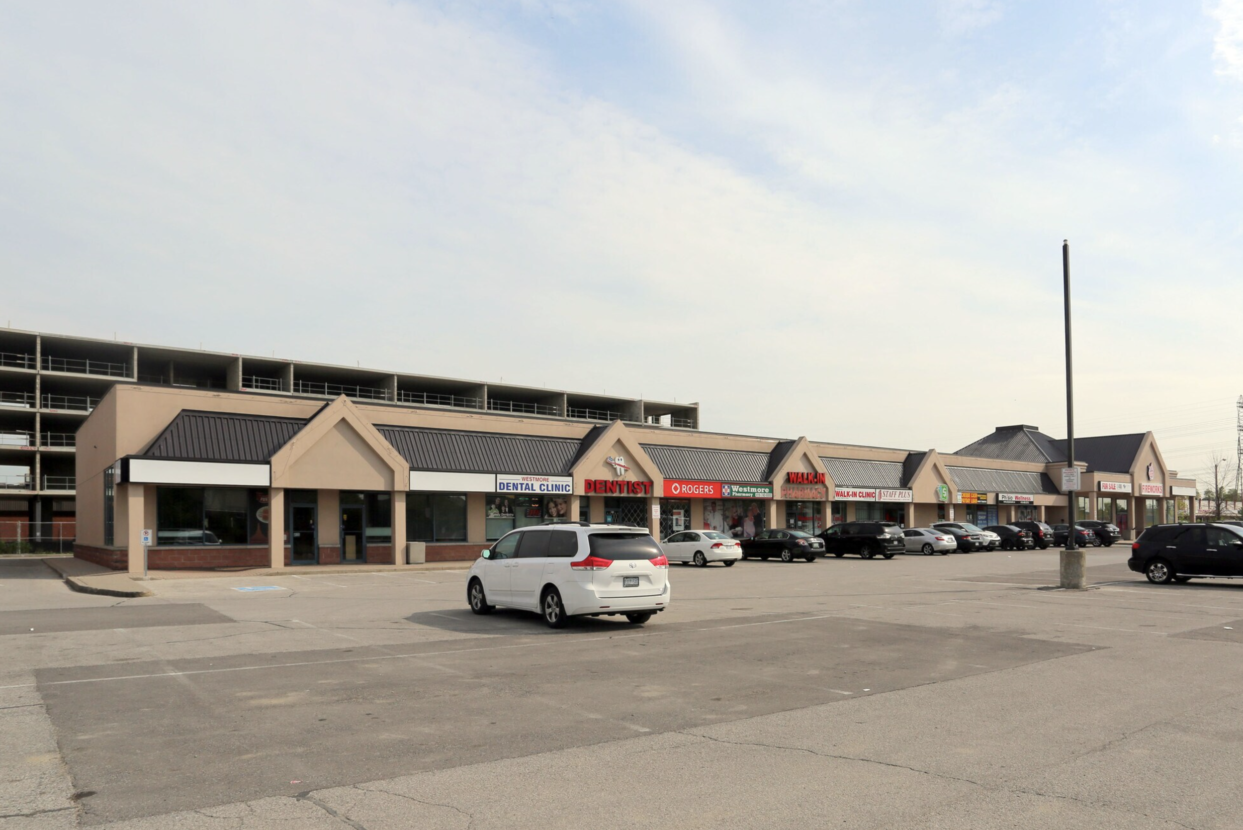 A strip mall with various businesses, including a dental clinic, pharmacy, and walk-in clinic, in front of a parking lot with several cars and a large empty space.
