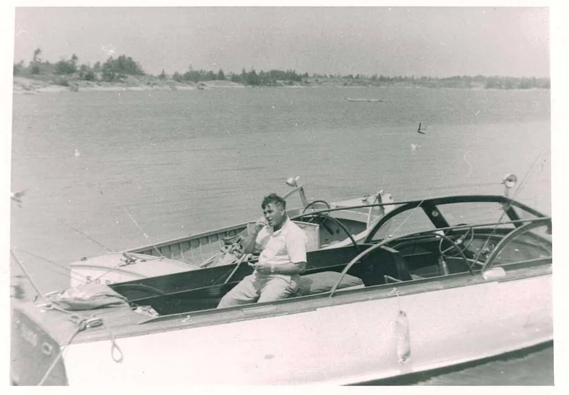 A young man sitting on the edge of a small boat with a larger boat in the background on a calm body of water.