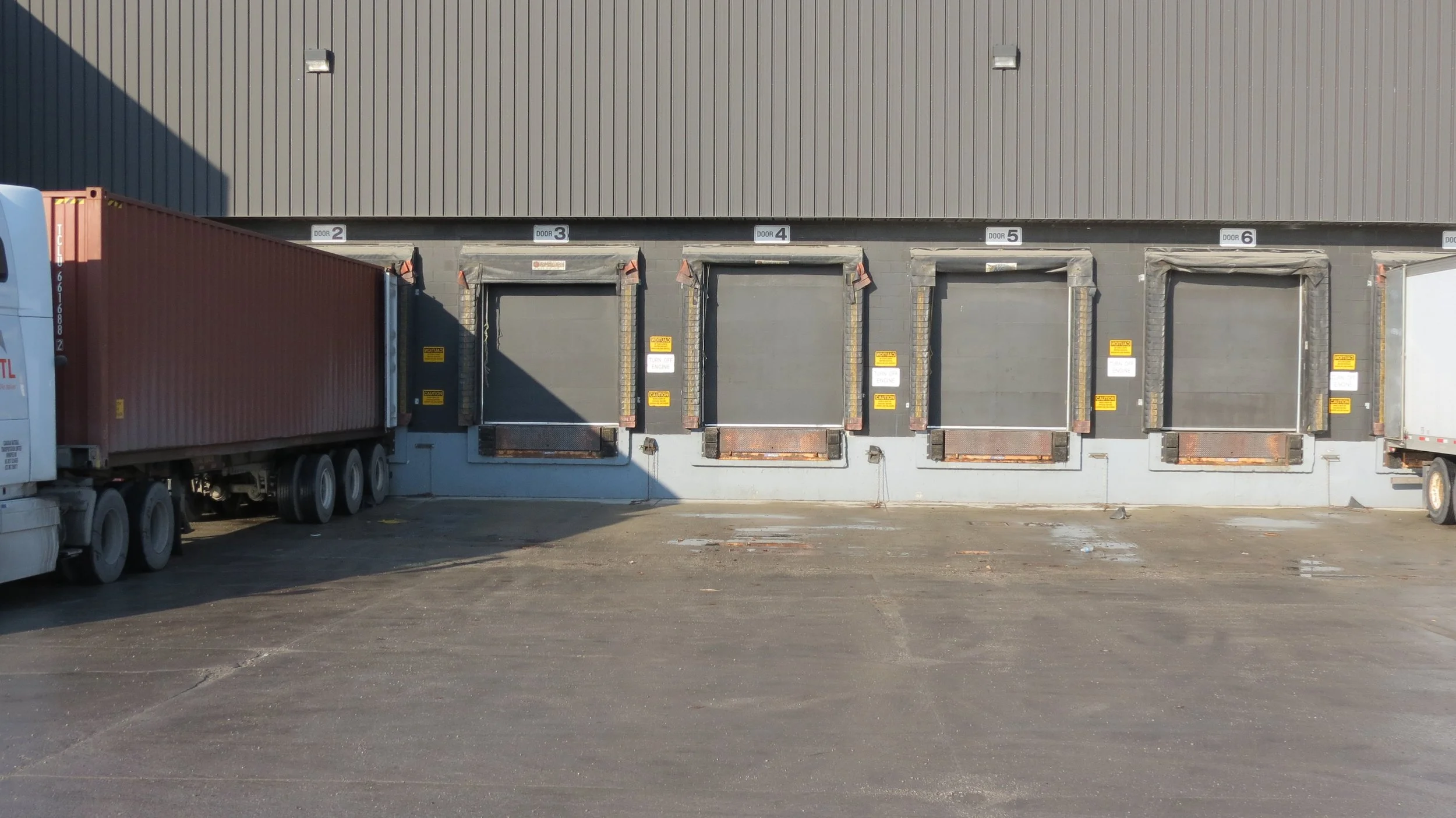 Loading dock area with five closed dock doors numbered 2 through 6, a red semi-truck trailer parked on the left, and a plain gray building. The ground is paved and has some patches of dirt and debris.