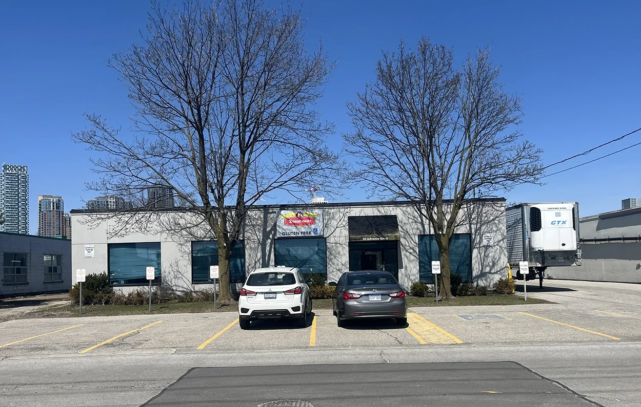 A white building with two leafless trees in front, three parking spaces, two cars parked, and a large truck on the right, under a clear blue sky.