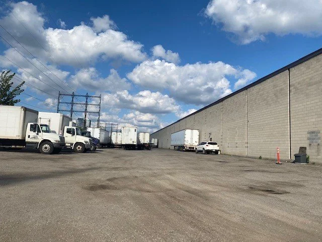 Parking lot with white trucks, a white sedan, a large beige warehouse wall, and a blue sky with clouds.