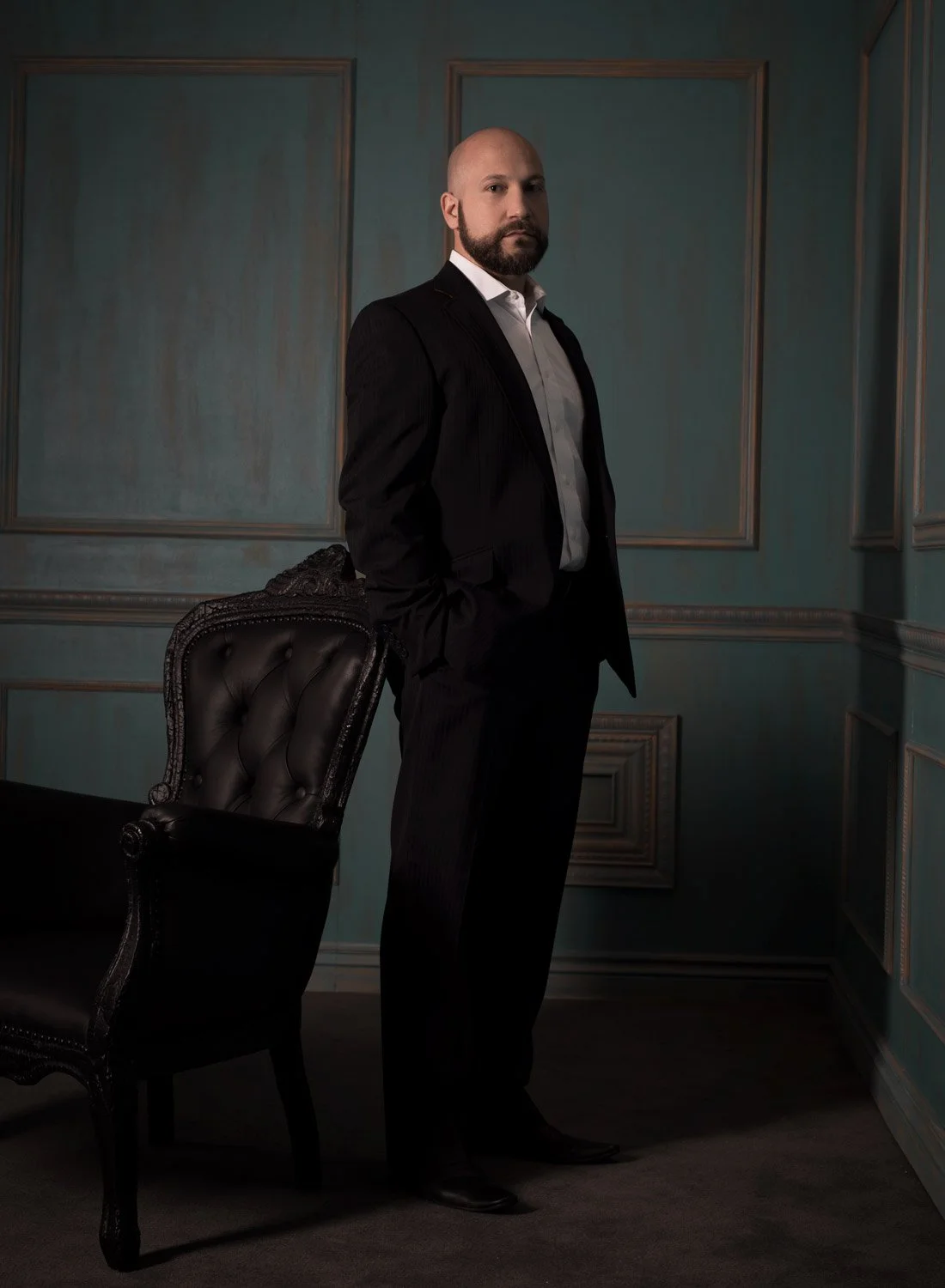 A man in a black suit standing in a dimly lit room with teal walls and decorative molding, with one hand in his pocket and leaning on an ornate black chair.
