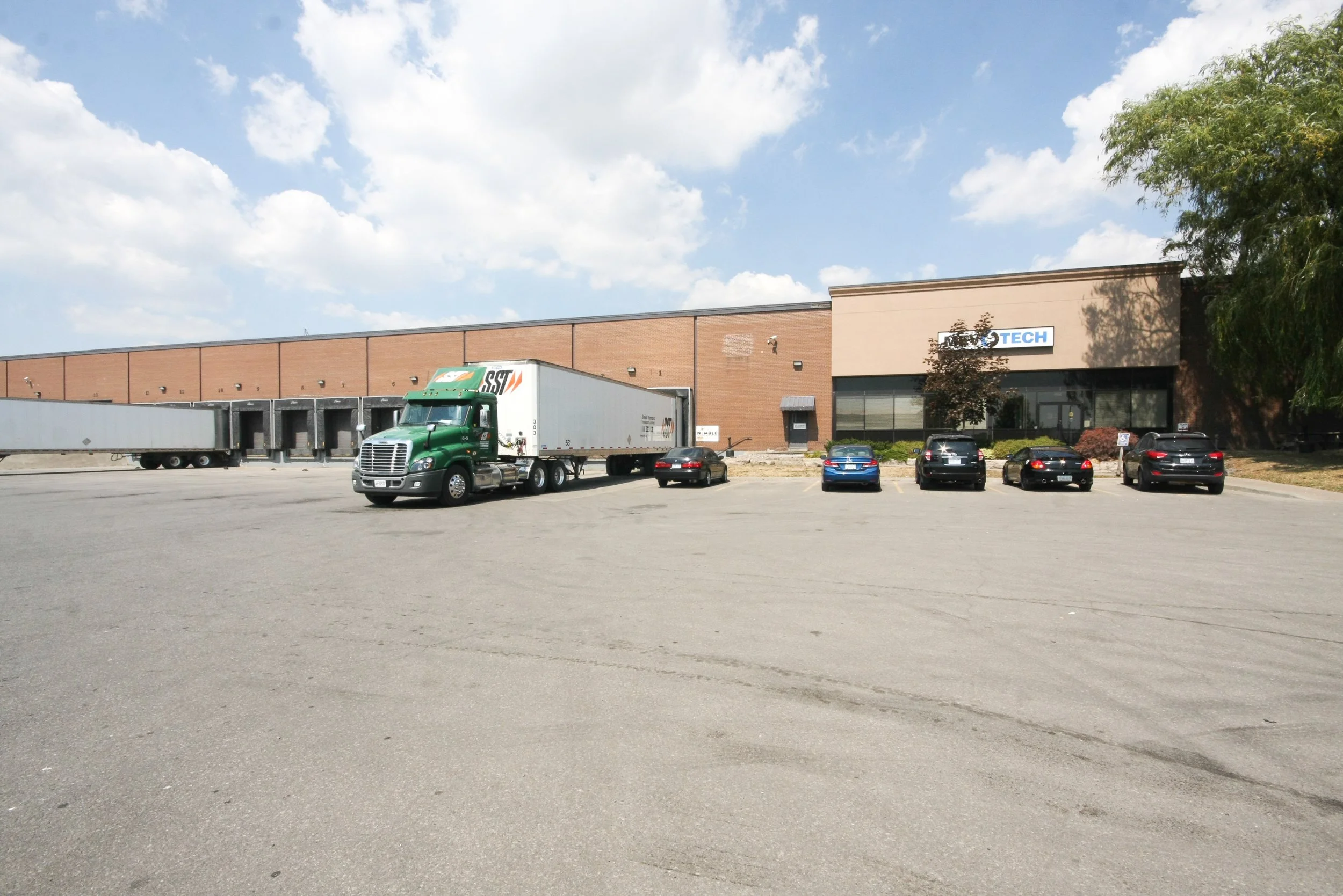 Parking lot in front of a large brick building with a sign that says 'TECH'. Several parked cars and a green semi-truck are visible under a partly cloudy sky.