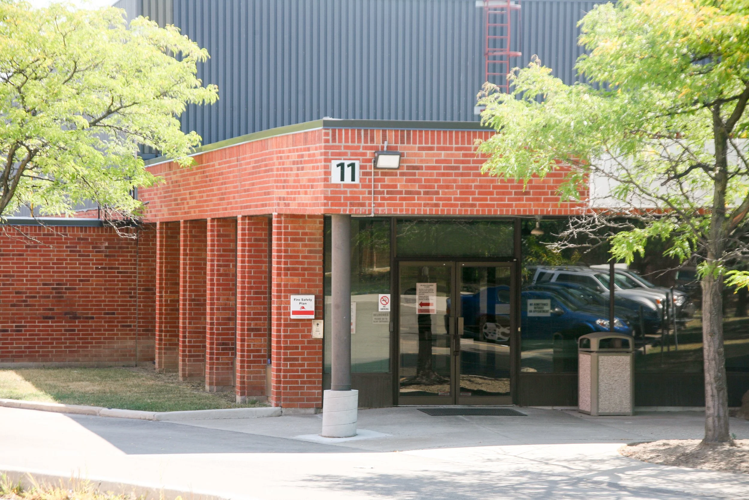 Street view of a brick building with trees on either side, a Belgian flag logo on the building, and a parking lot in front. The building has a sign that reads 'Fire Safety Plan' near the entrance.