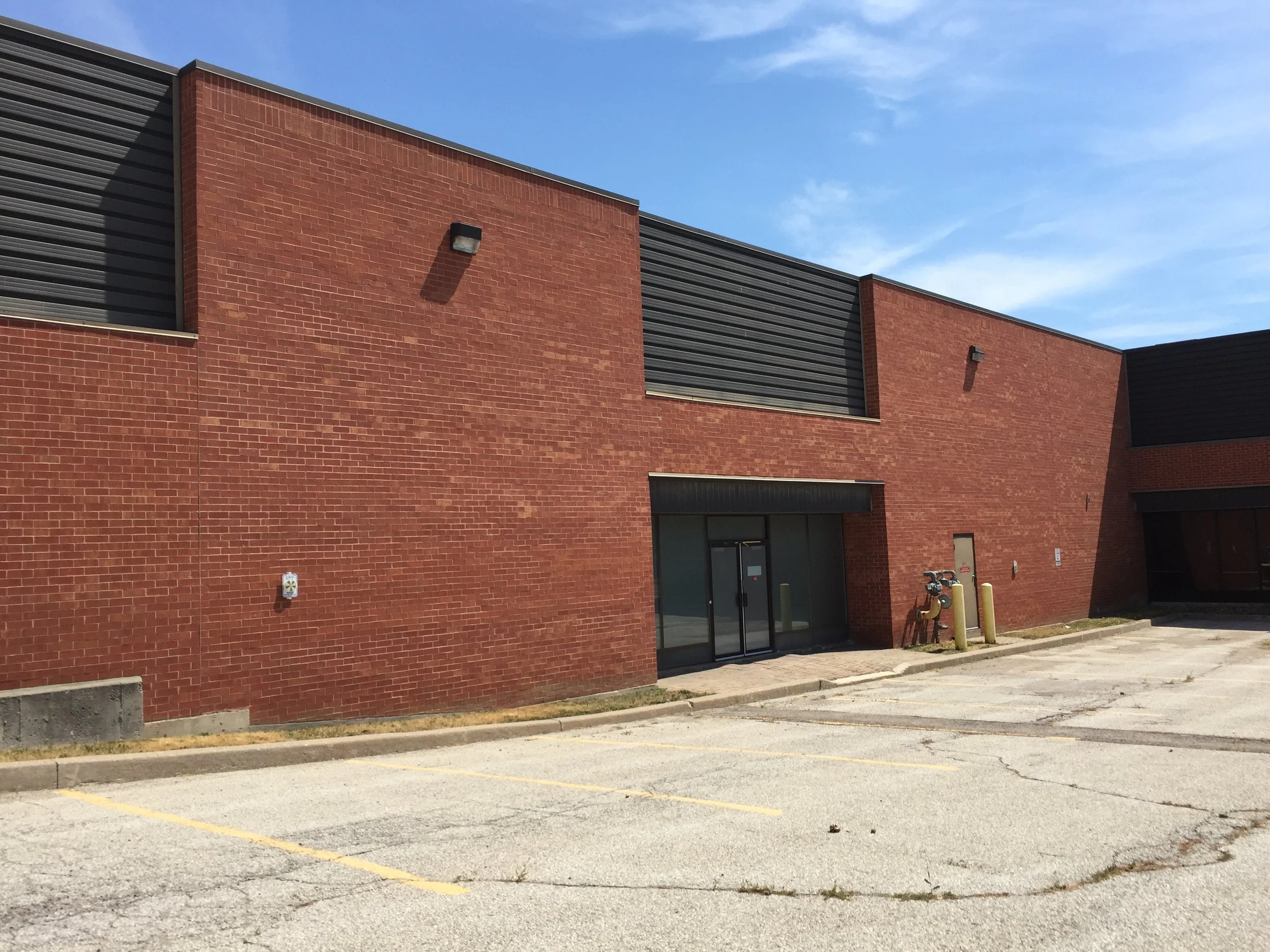 Exterior view of a modern brick building with dark gray siding, black door, and small windows, under a blue sky with some wispy clouds. There are sidewalks and yellow parking lot lines in the foreground.