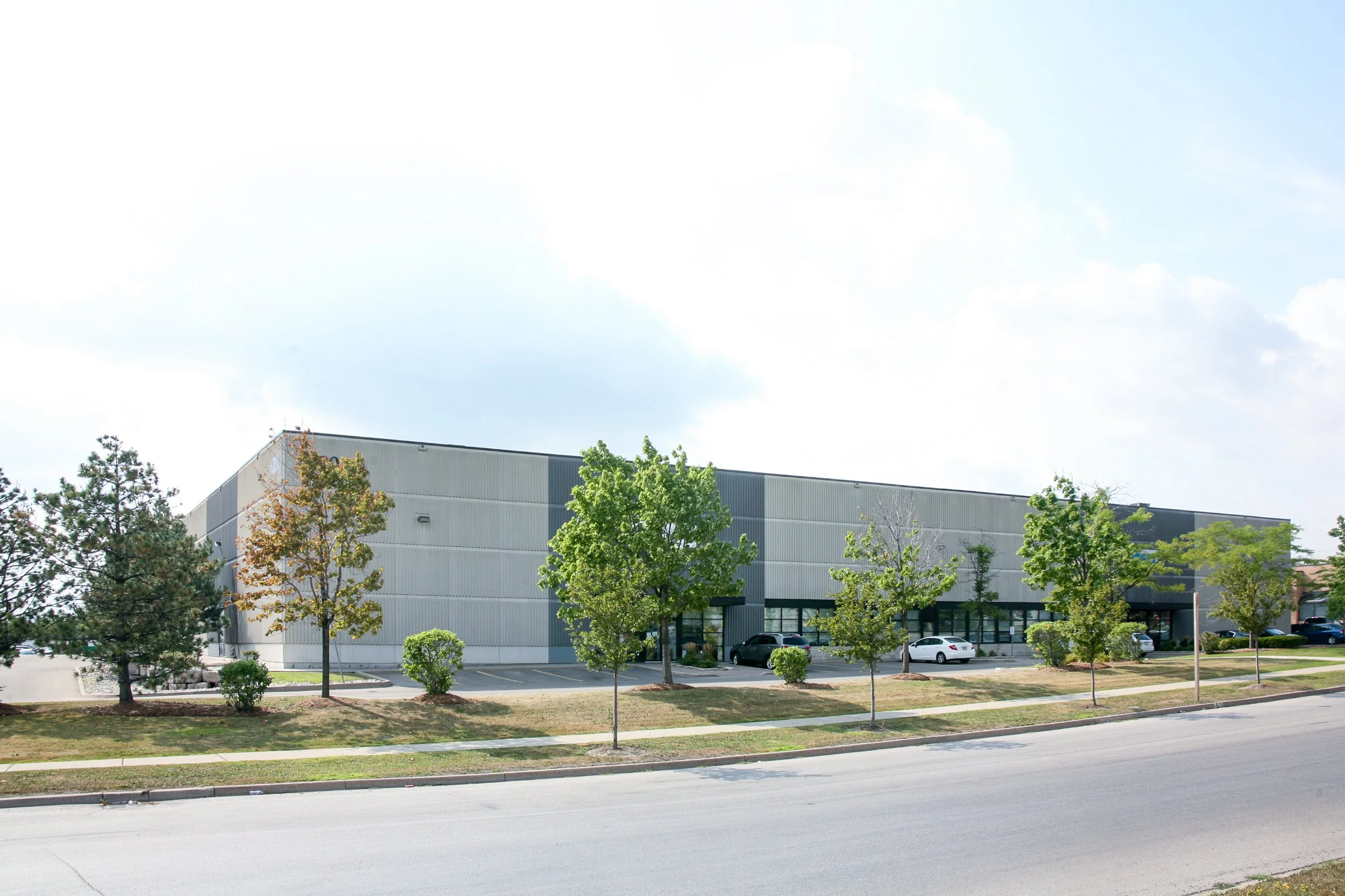 A large industrial building with a gray exterior, surrounded by a parking lot with several parked cars and a row of green trees in the foreground, under a partly cloudy sky.