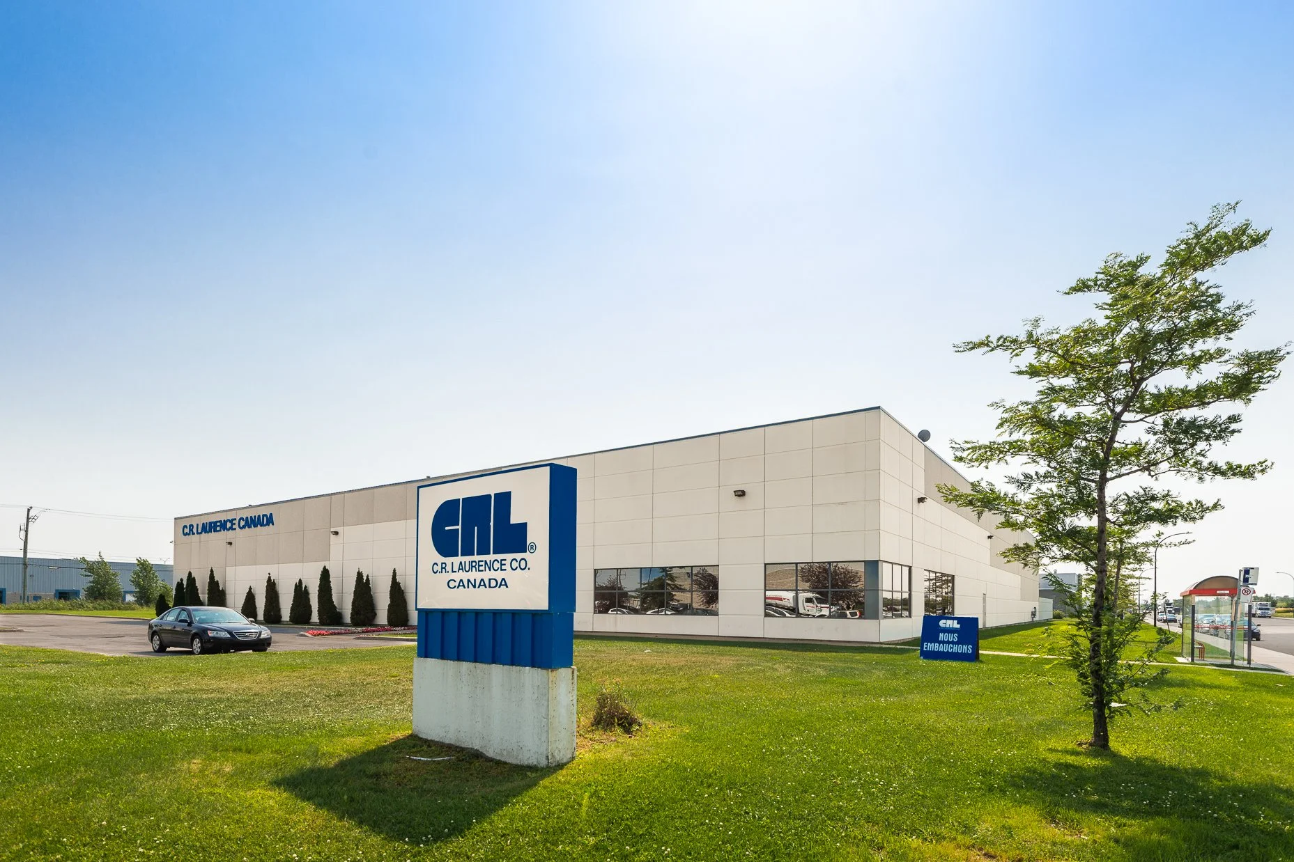 The exterior of a modern commercial building with a sign that reads "C.R. Laurence Co. Canada." The building is white with large windows, surrounded by a well-manicured lawn with a few trees, and a parking lot with a car sitting in front. The sky is clear with bright sunlight.