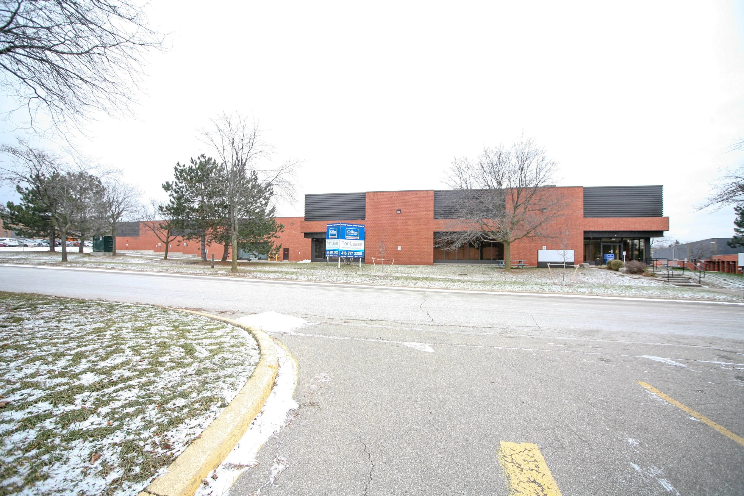 A brick building with a black stripe on top and blue signs that say 'For Lease,' situated in a parking lot with some snow on the ground and leafless trees around.