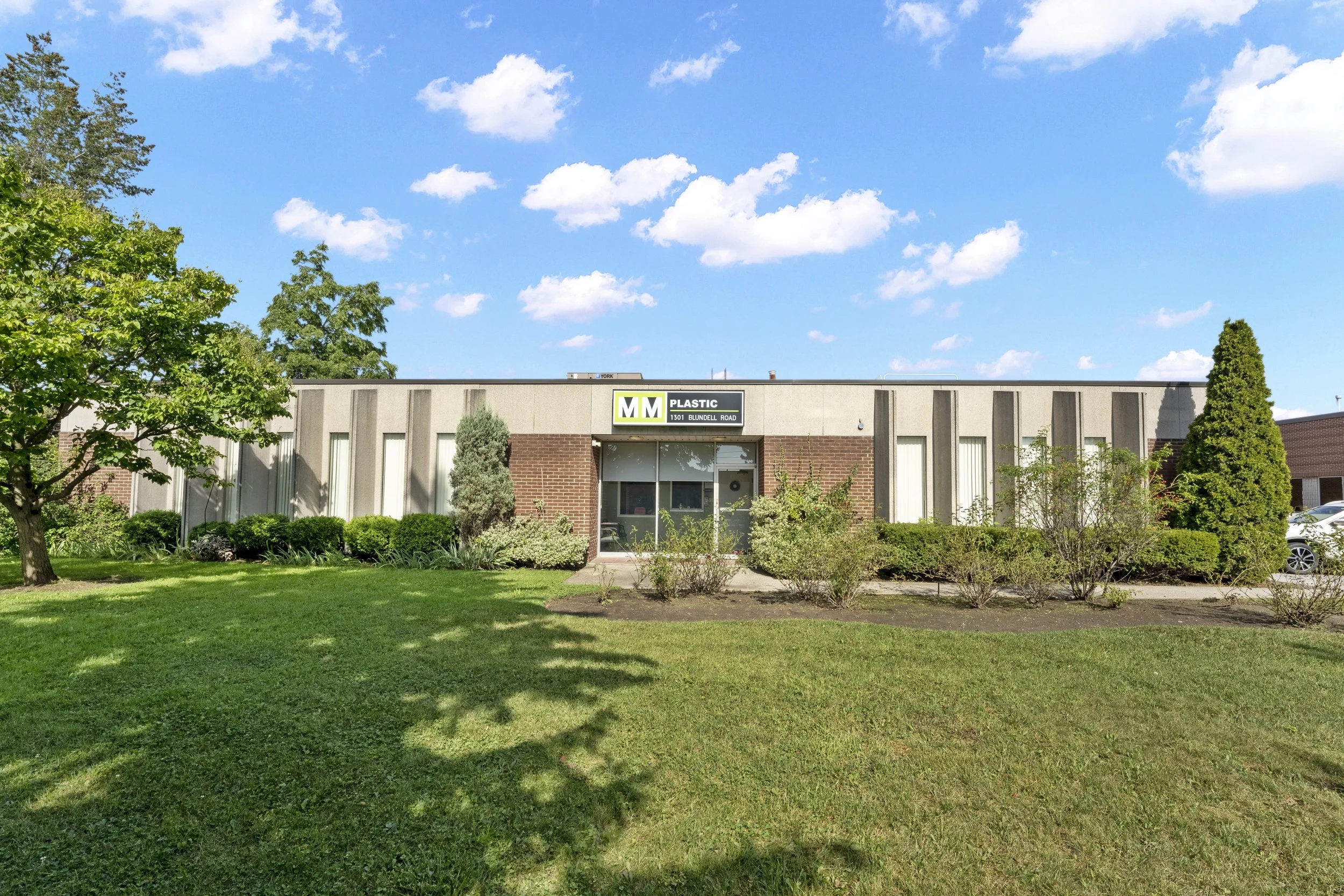 A single-story commercial building with a sign that reads 'MM Plastic' and an address 1301 Blundell Road, surrounded by greenery, under a blue sky with scattered clouds.